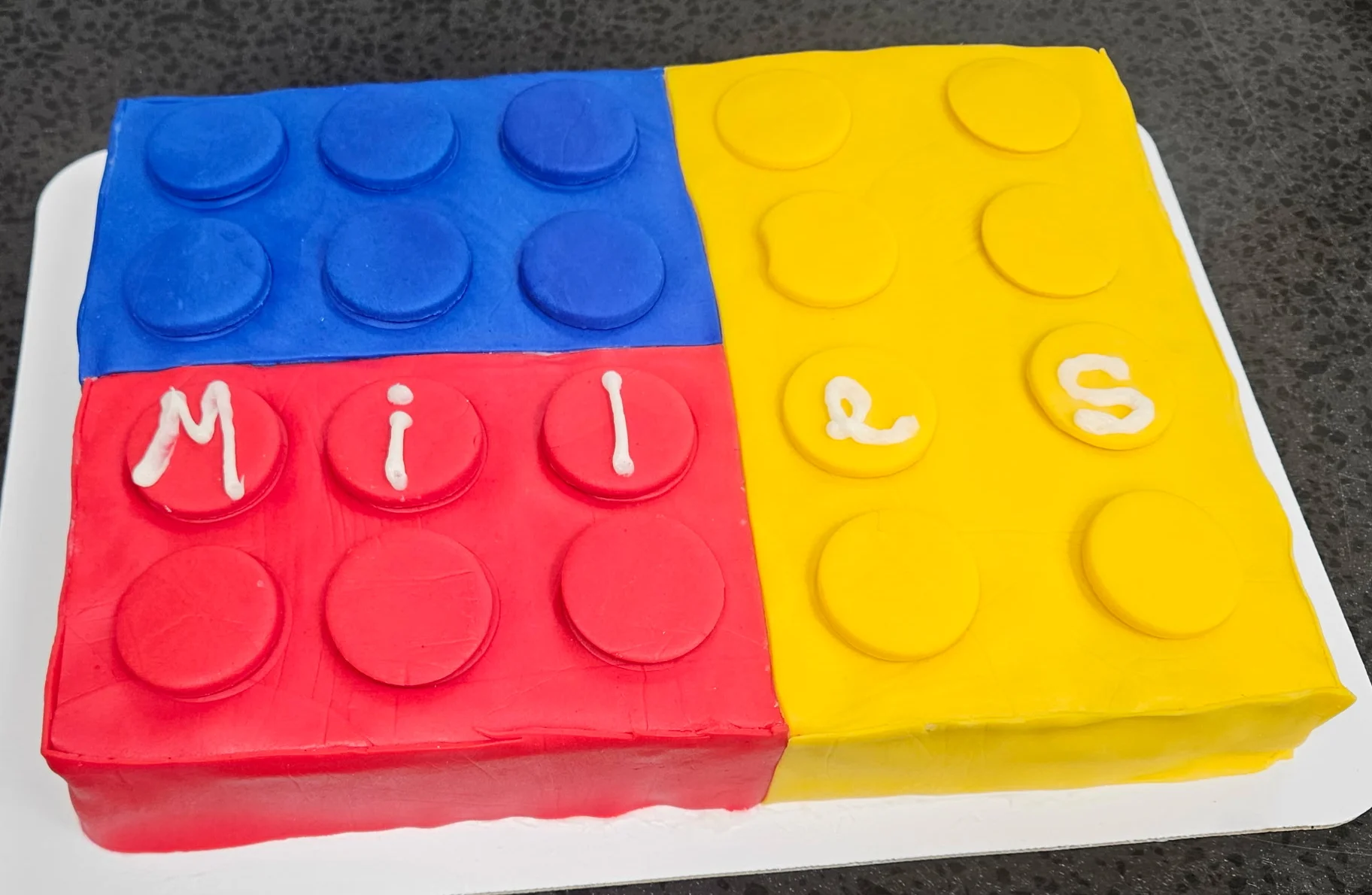 Sheet cake covered in primary color fondant to look like Lego bricks and the name "Miles" iced in the center.