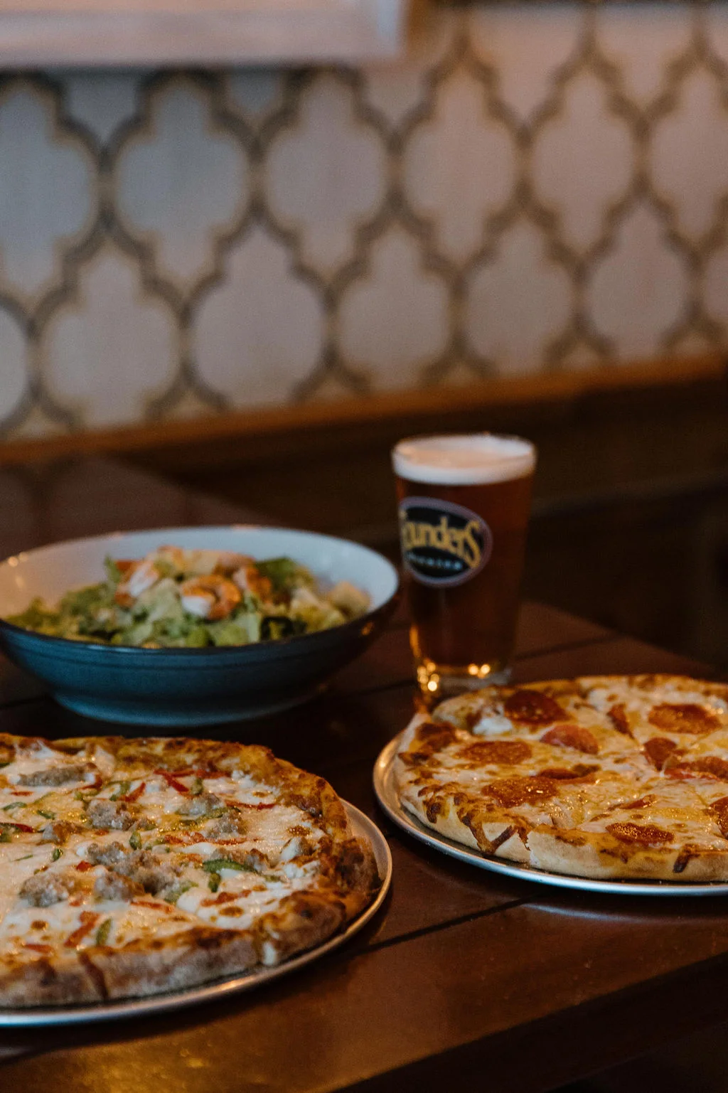 A table featuring two pizzas, a bowl of salad, and a pint of beer. The pizzas showcase a variety of toppings on crispy crust.