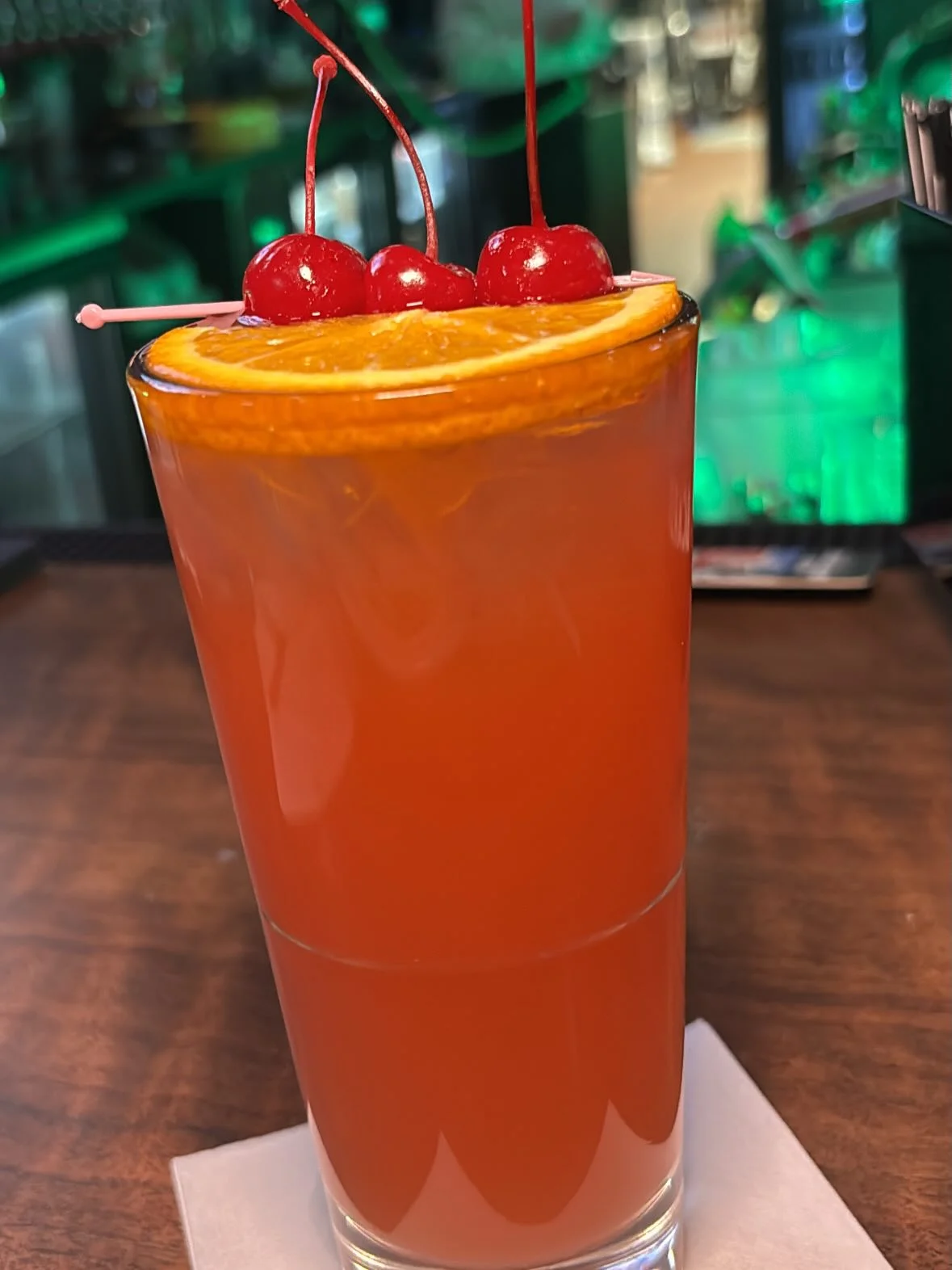 A vibrant cocktail served in a tall glass, garnished with cherries and an orange slice.