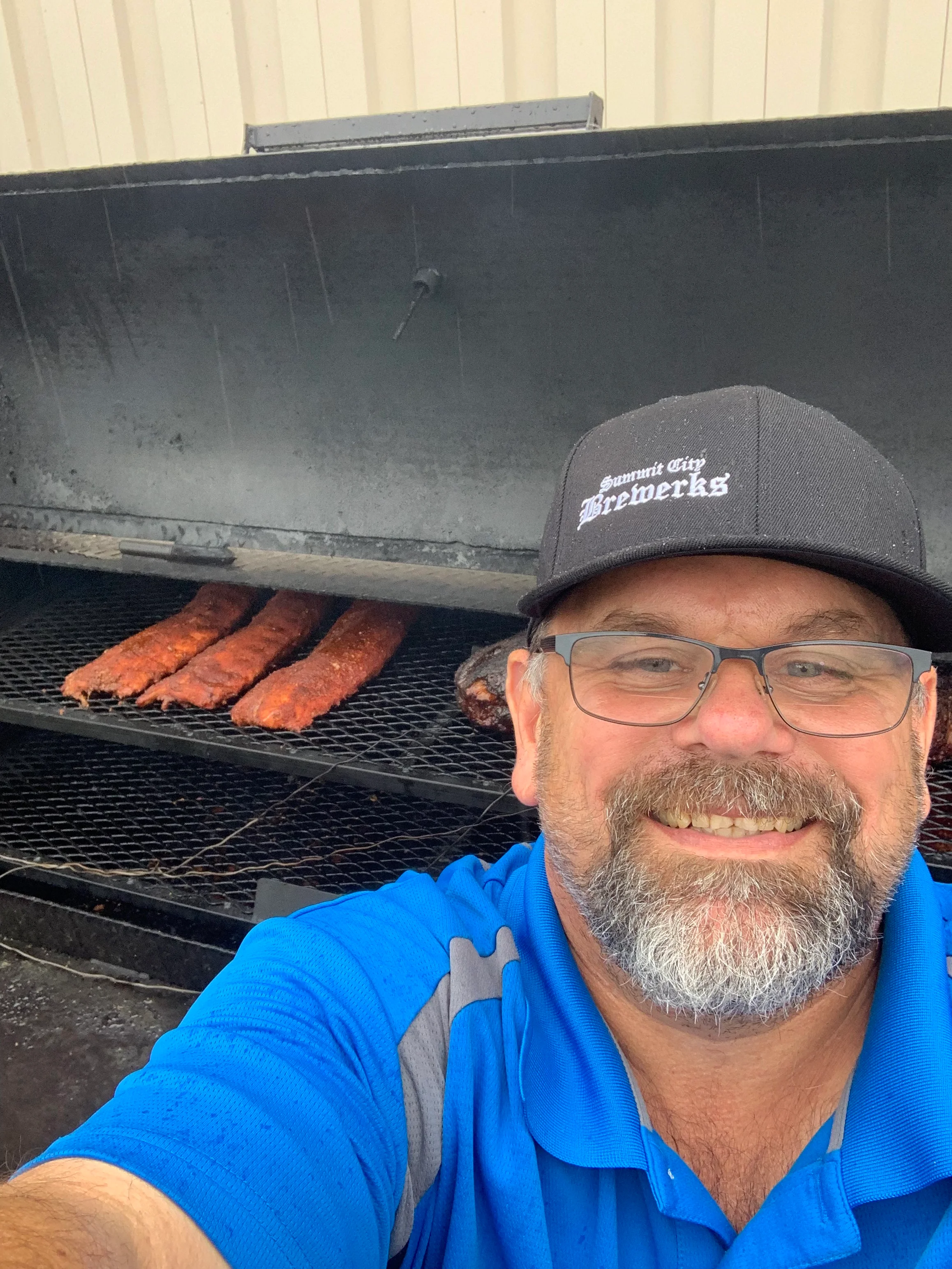 A smiling individual taking a selfie in front of a large smoker with partially cooked ribs inside.