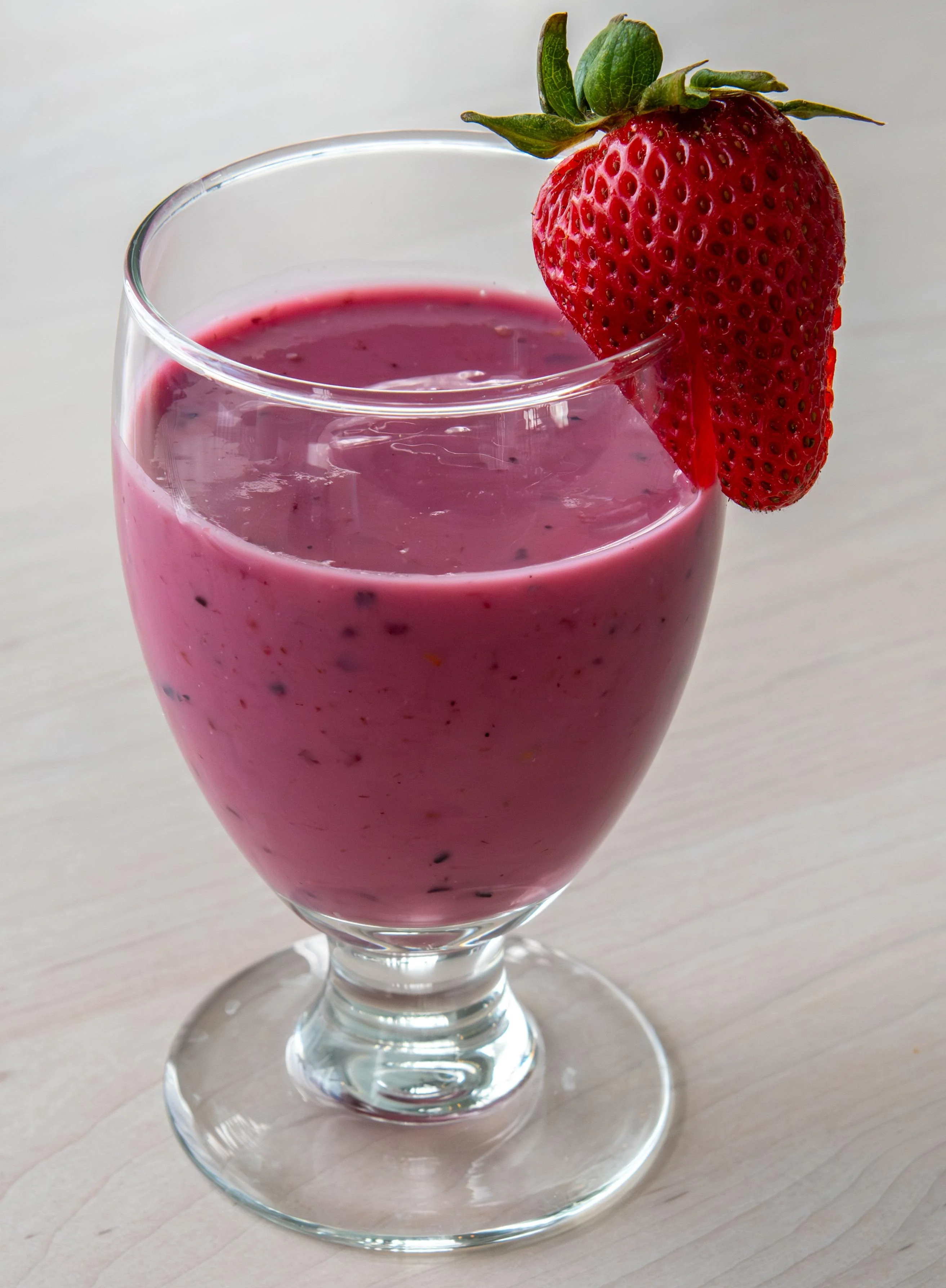 A glass of pink smoothie topped with a fresh strawberry. The smoothie appears creamy and is presented in a tall glass.