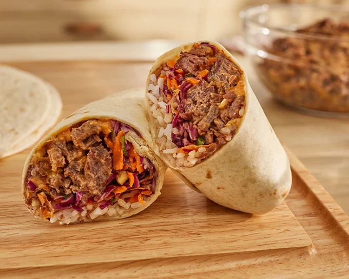 A grilled burrito cut in half, revealing a filling of beef, rice, shredded vegetables, and sauce, served on a wooden board.