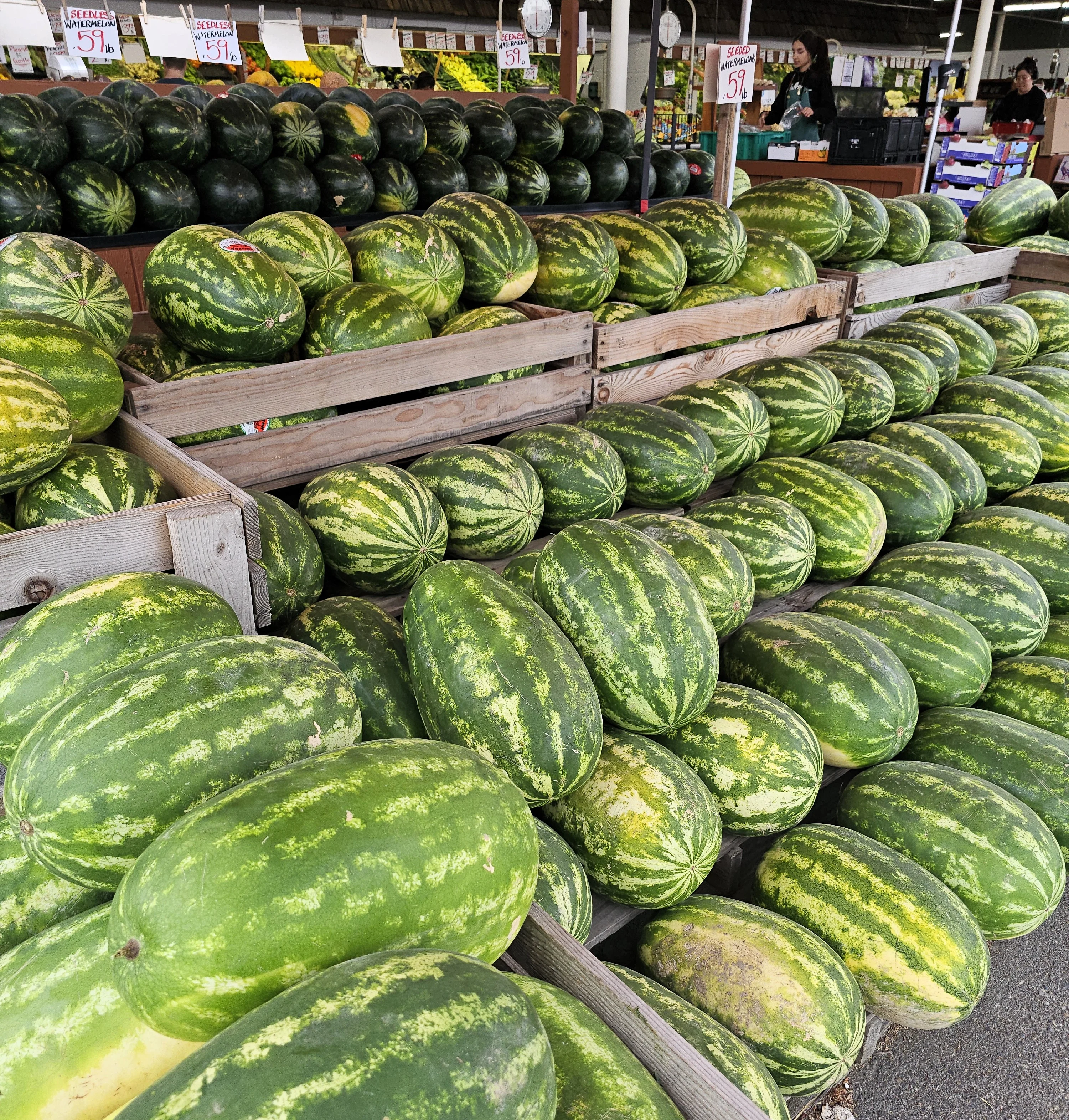 An outdoor market stall named 'Country Farms' featuring a variety of fruits and vegetables, with watermelons prominently displayed under large umbrellas.