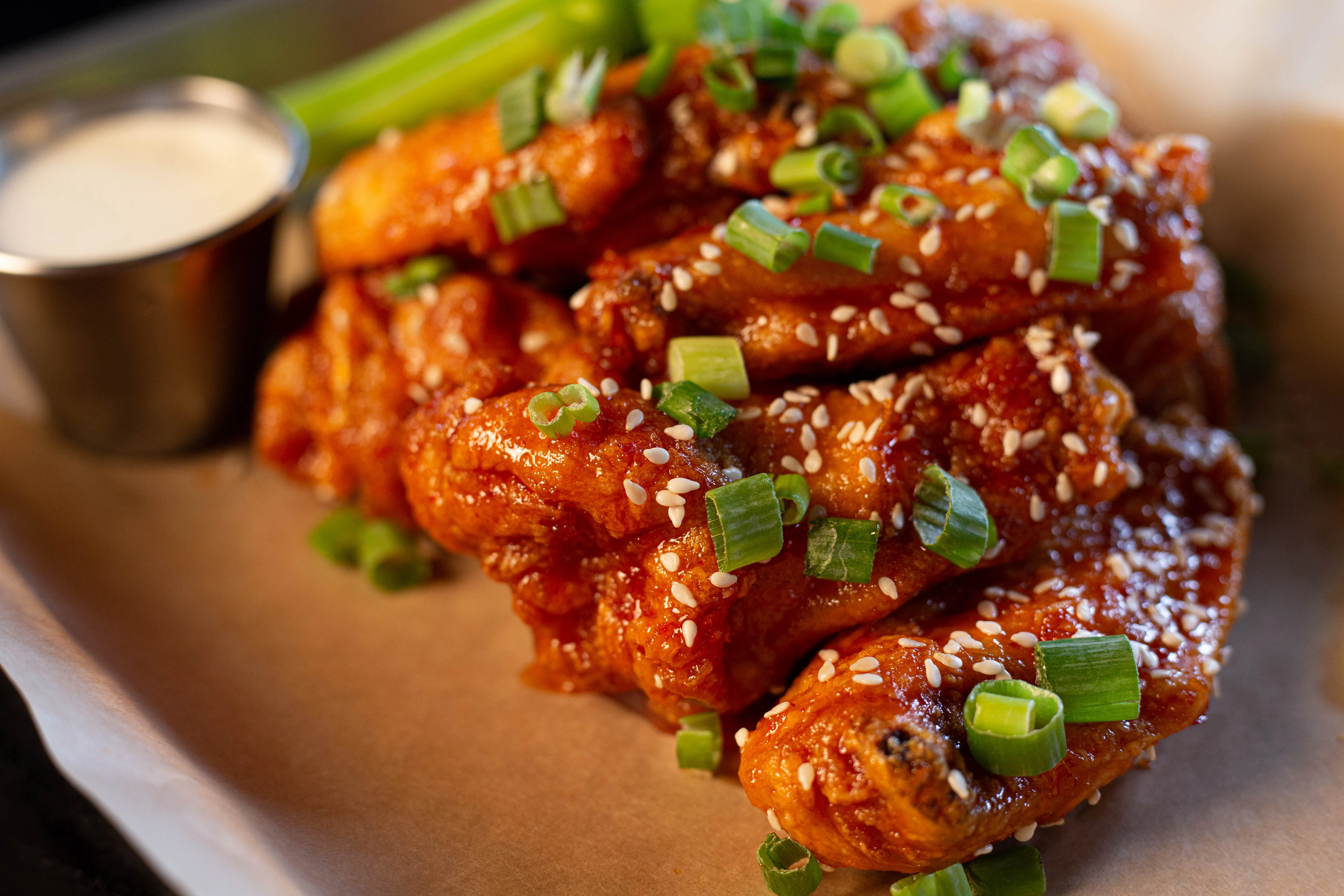 A close-up of glazed chicken wings garnished with sesame seeds and green onions, served with a side of creamy dipping sauce.