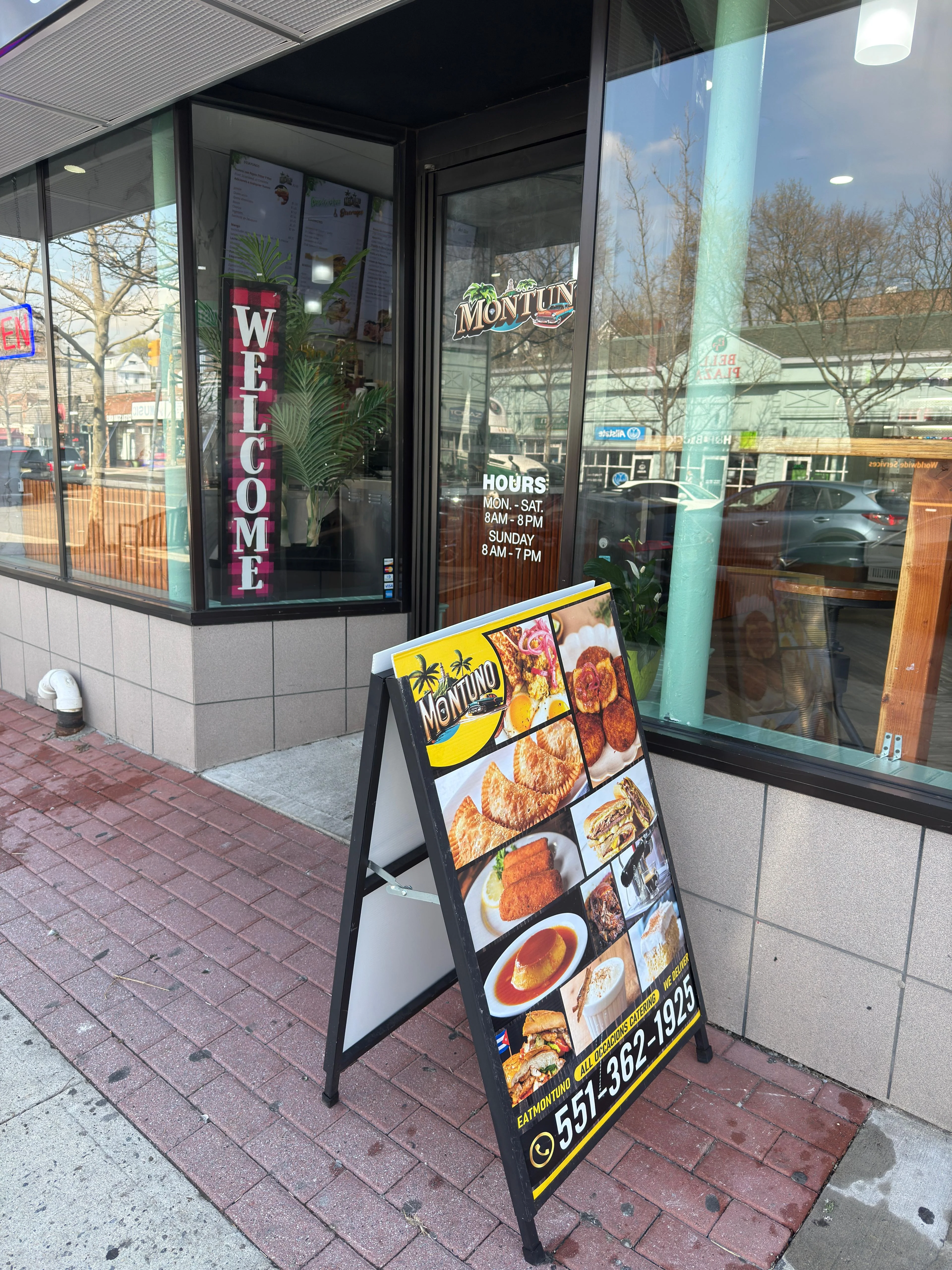 The storefront of 'Montuno' restaurant with a glass door and window displaying a large 'WELCOME' sign. The door lists the restaurant's operating hours: Monday to Saturday, 8 AM to 8 PM, and Sunday, 8 AM to 7 PM. In front of the restaurant, there is a sandwich board with the restaurant's name, menu highlights, a contact number (551-362-1925), and the social media handle '@EATMONTUNO.' The sign also mentions that all orders can be placed online.