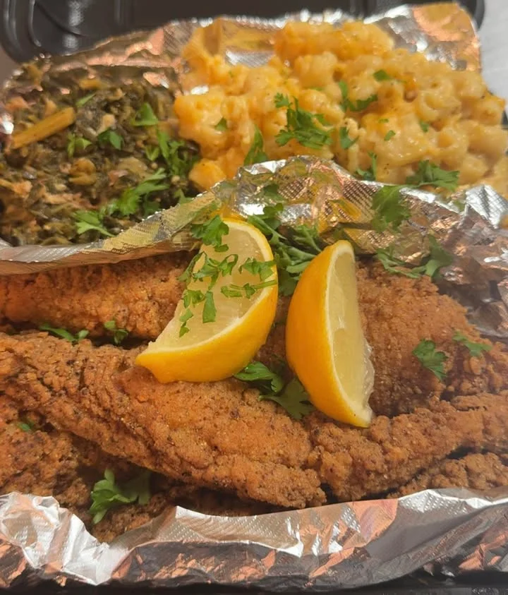 A takeaway food container filled with fried catfish, macaroni and cheese, and collard greens, garnished with lemon and herbs.