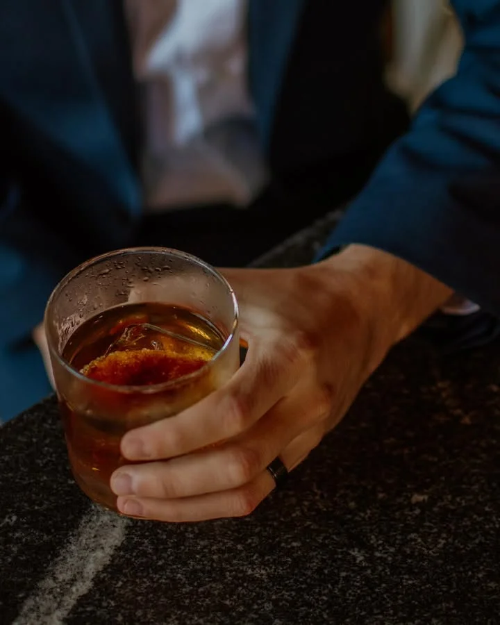 A hand holding a glass of dark-colored beverage with ice, resting on a textured surface.