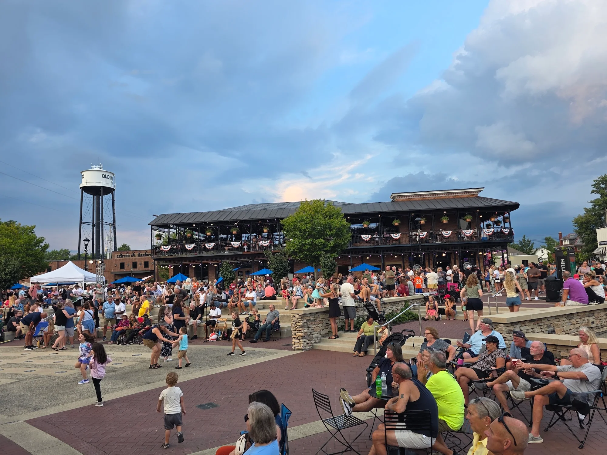A lively outdoor event with a large crowd gathered around a performance area and food tents.