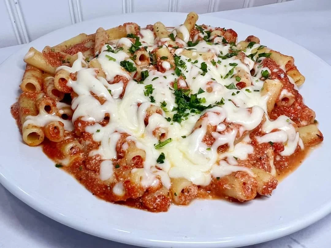 A serving of baked pasta topped with melted cheese and parsley, served in a white plate with a rich tomato sauce.