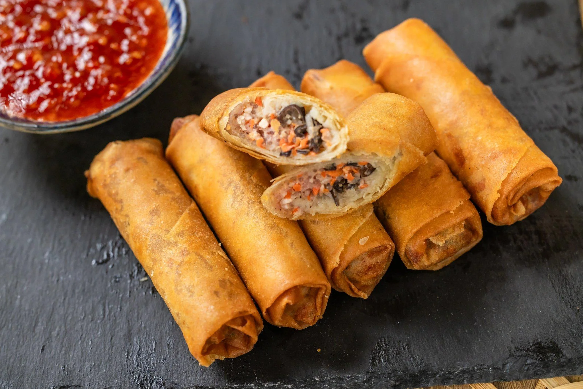 Several golden-brown spring rolls on a slate platter with one cut in half to reveal the filling, served alongside a dipping sauce.
