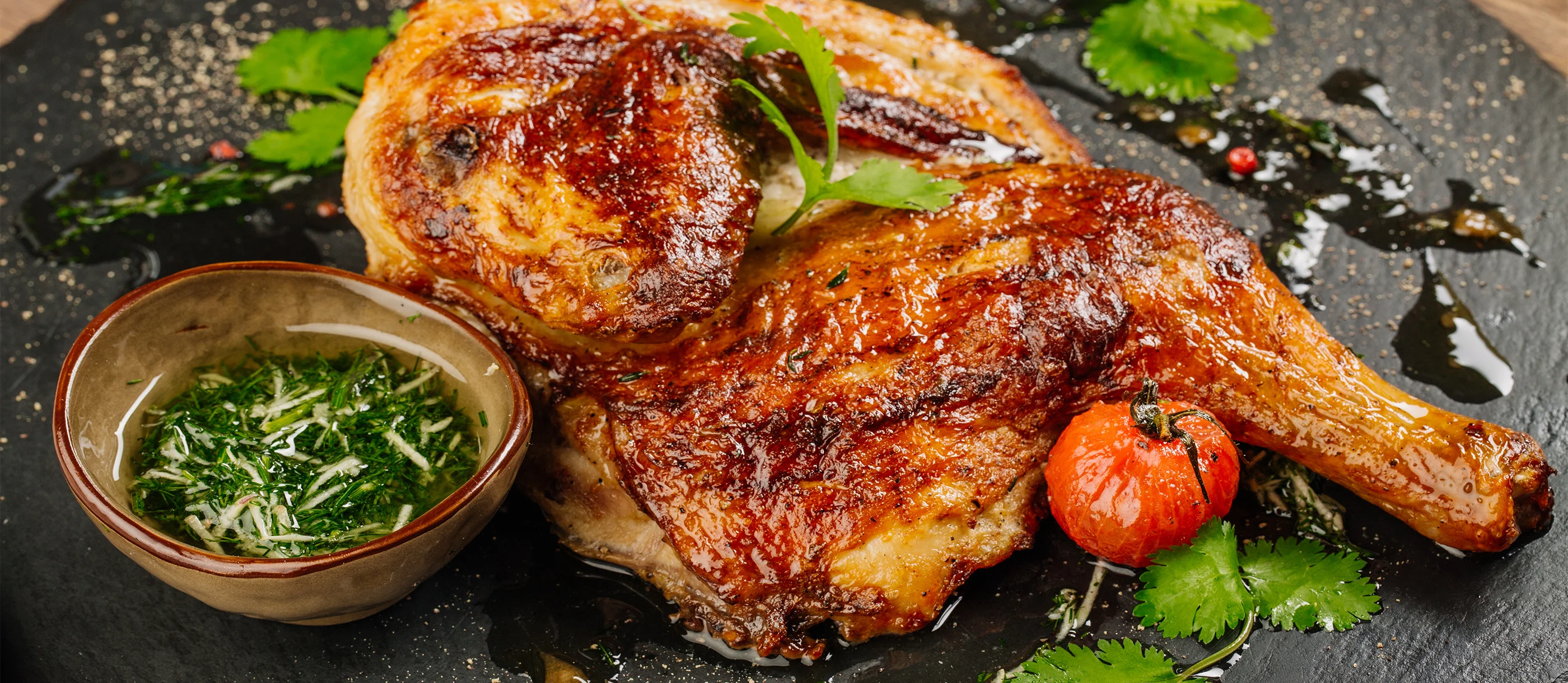A golden-brown chicken with a side of sauce, presented on a slate plate.