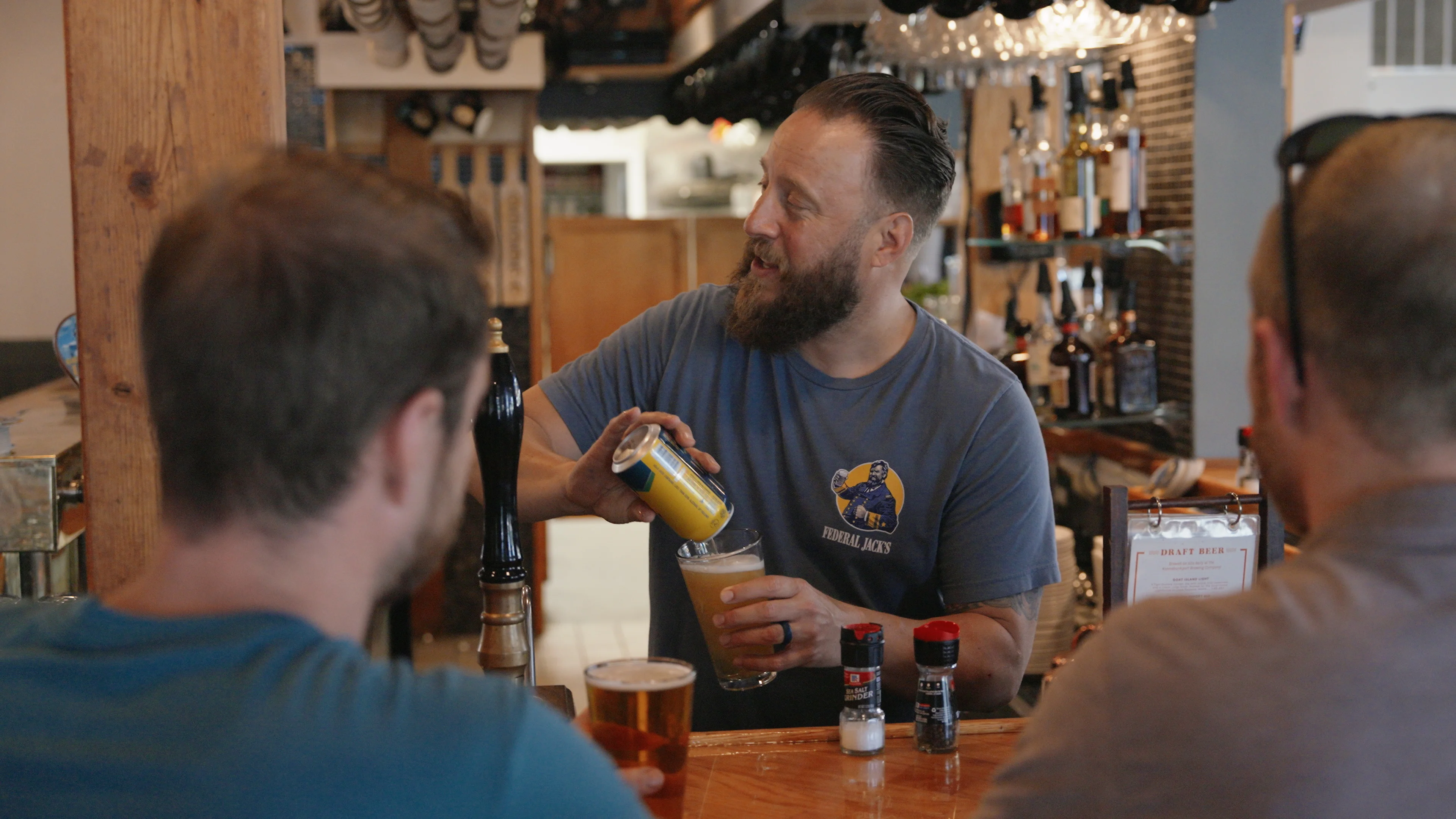 A bartender pours a drink from a can into a glass, while customers enjoy their drinks at the bar.