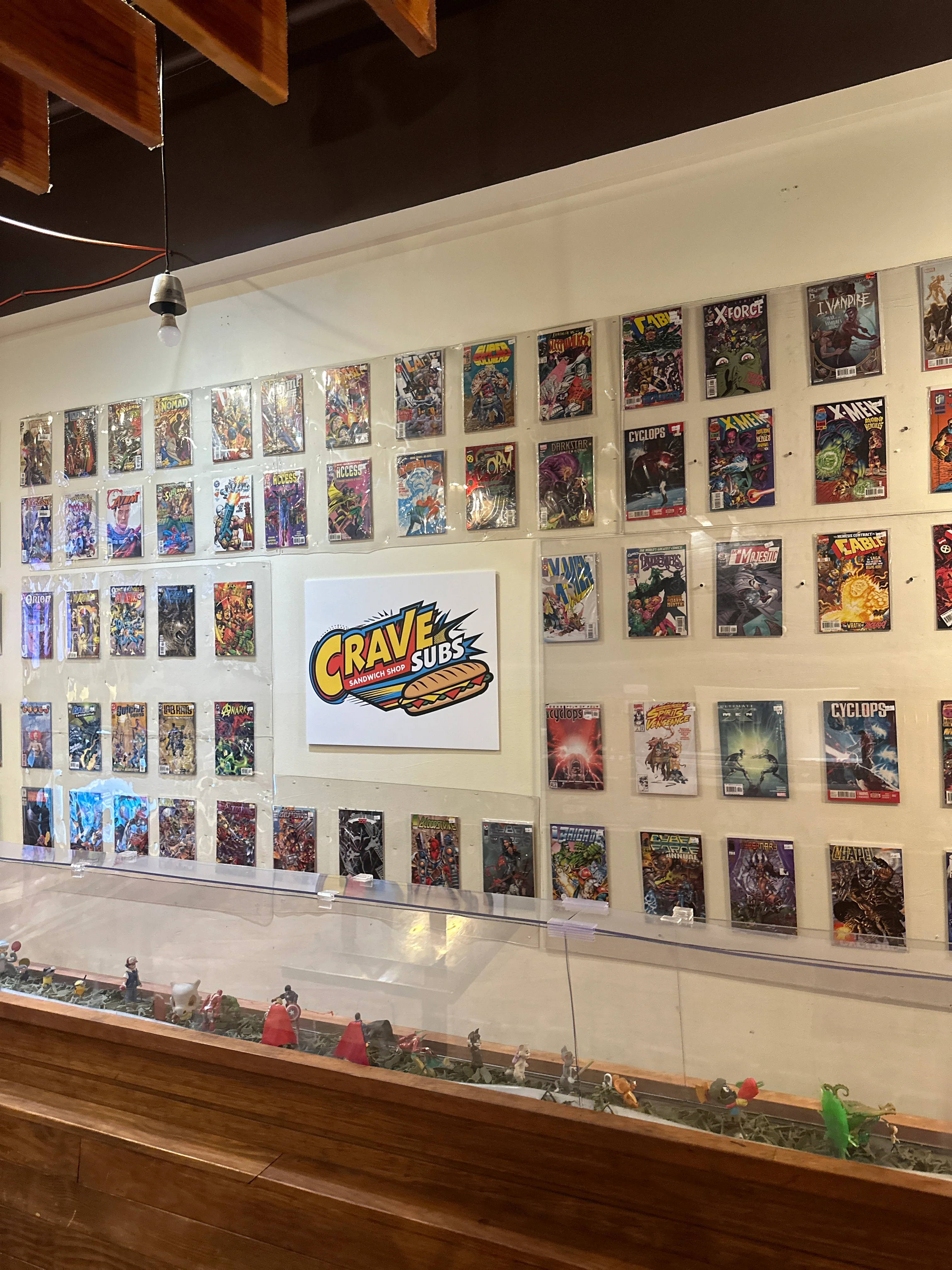 A display wall filled with various comic book covers, showcasing vibrant and colorful artwork. The wall includes a prominent sign for 'Crave Subs' in the center.