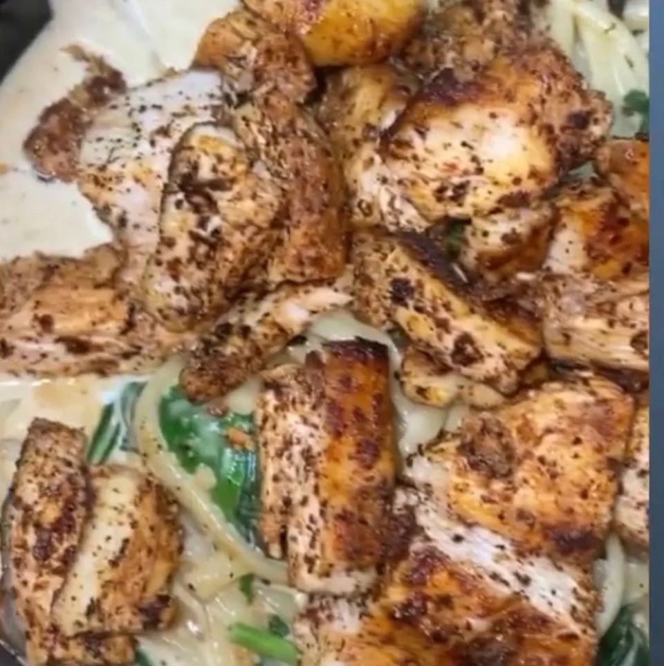 A plate of seasoned grilled chicken over a bed of fettuccine noodles with a creamy sauce and spinach.