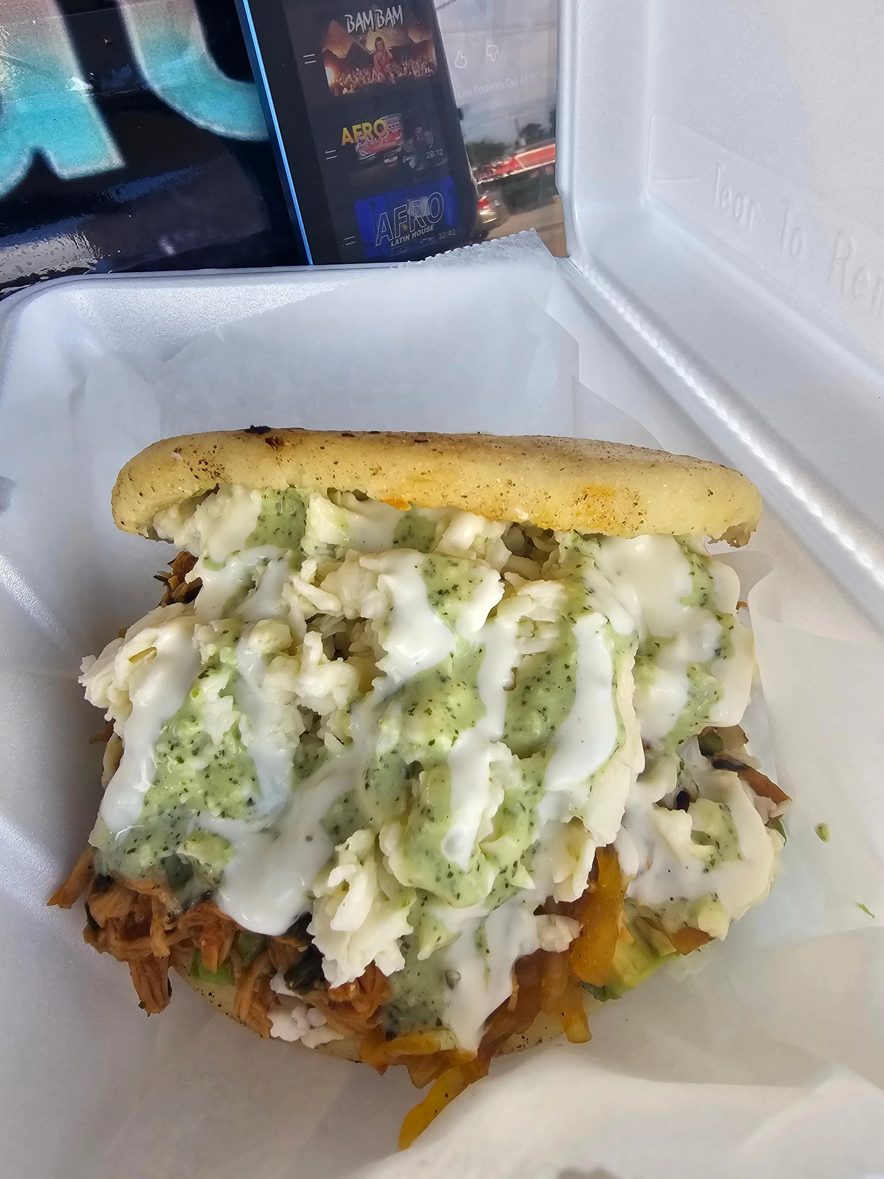 A stuffed arepa filled with shredded meat, cheese, rice, and drizzled with green and white sauces, presented in a takeout container.