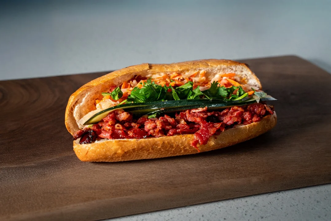A sandwich filled with a mix of meat, pickled vegetables, and fresh herbs, served in a toasted baguette.