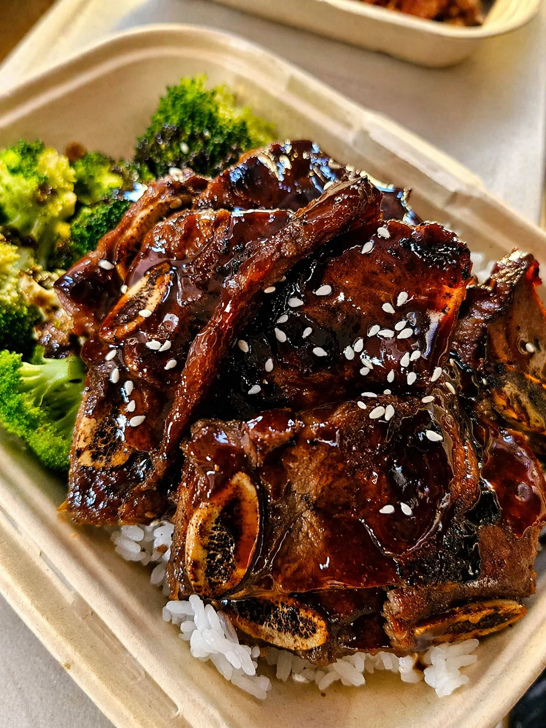 A serving of tender ribs glazed with a sweet sauce, served over rice with steamed broccoli and sesame seeds for garnish.