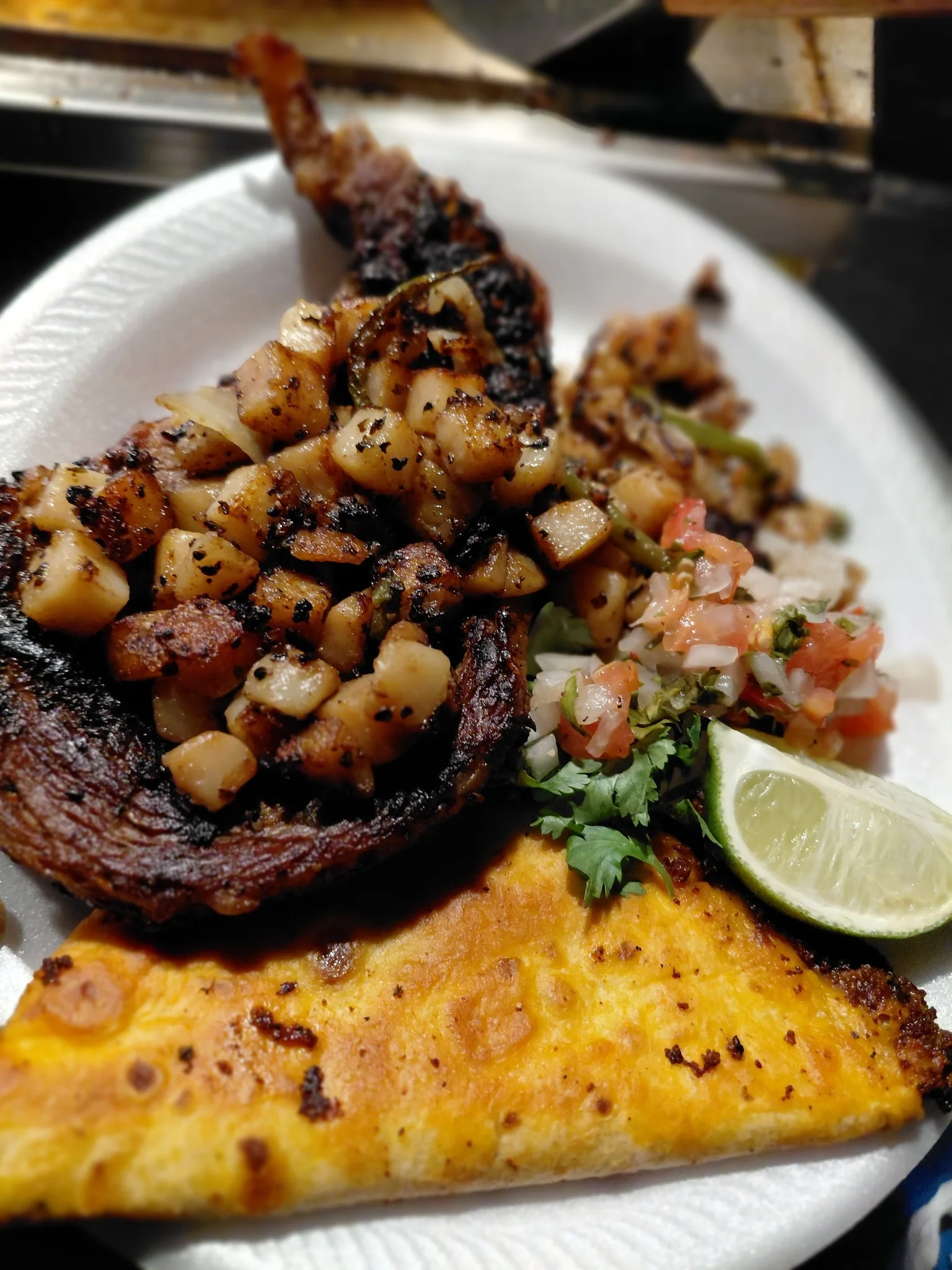 A serving of grilled meat topped with diced onions and served with a side of pico de gallo, lime, and a slice of fried cheese on a disposable plate.
