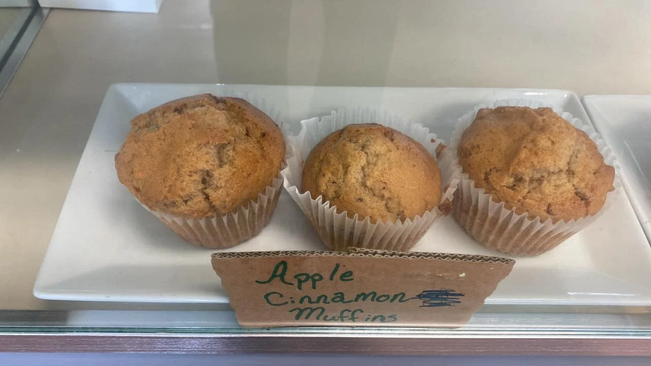 Three apple cinnamon muffins showcased in a bakery case, labeled accordingly.