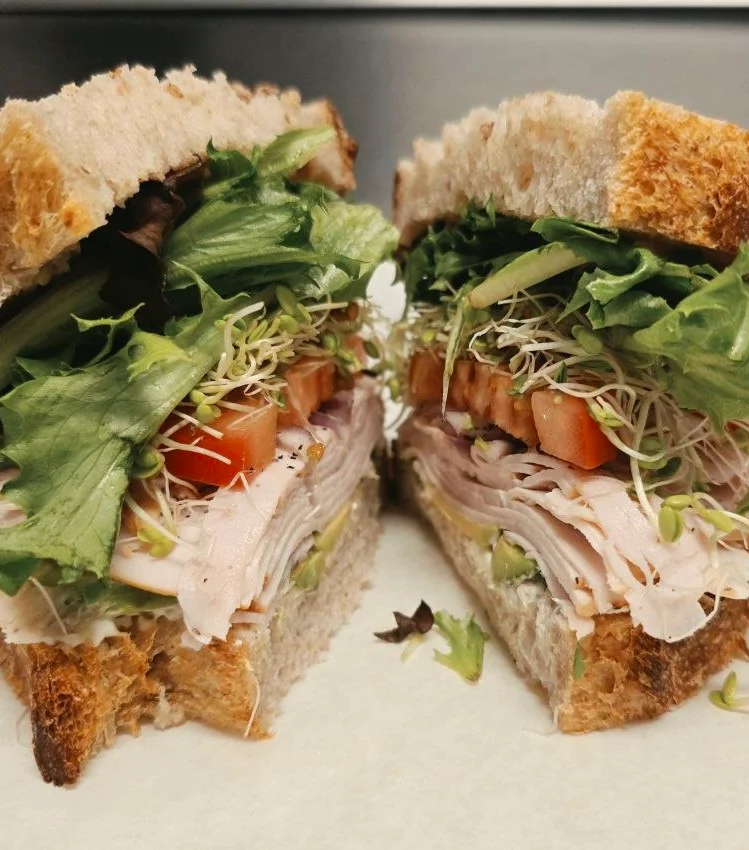A freshly made sandwich held in a hand, filled with various layers of meat and vegetables, set against a green backdrop.