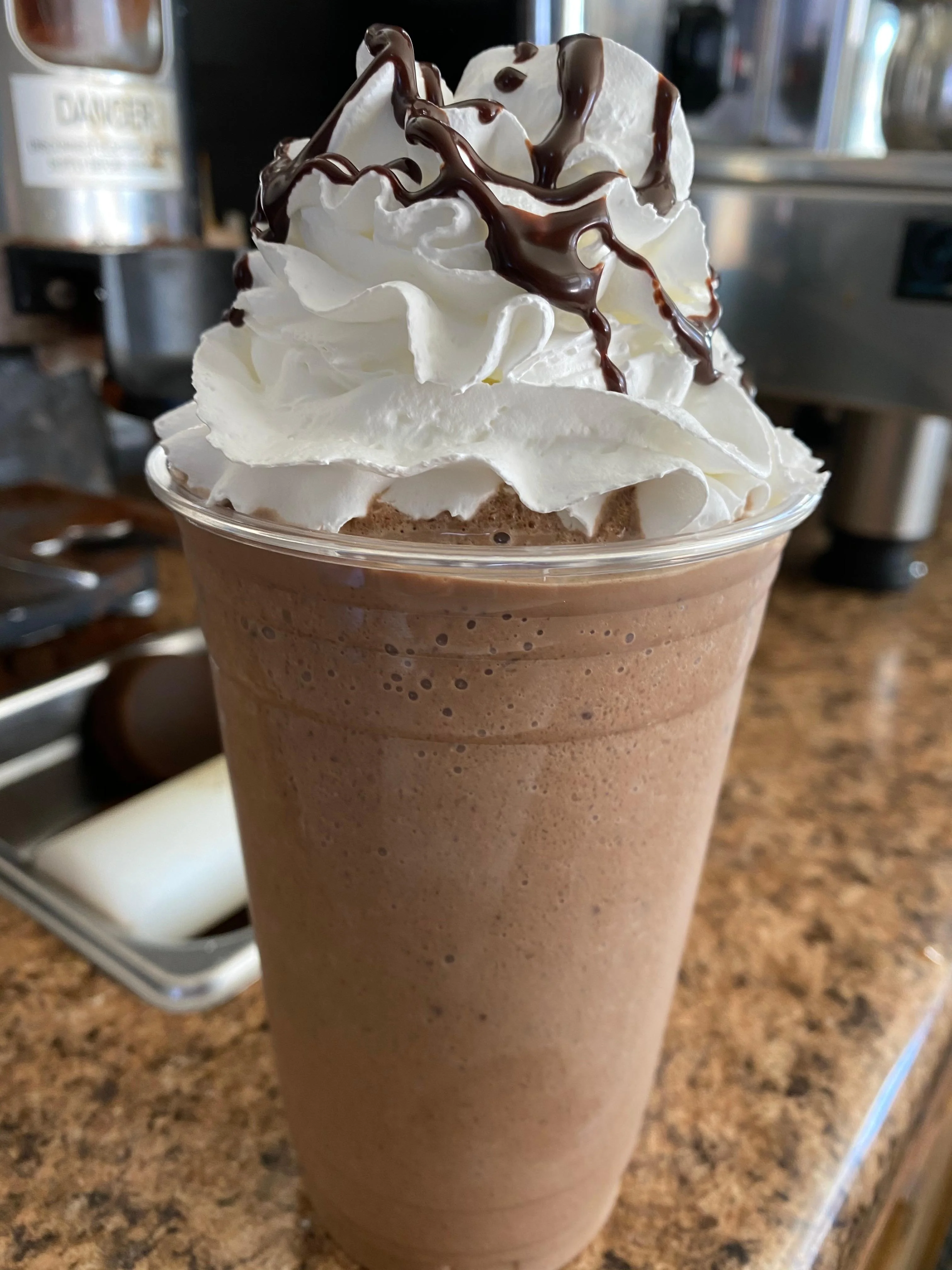 A chocolate milkshake topped with whipped cream and chocolate drizzle, served in a clear cup.