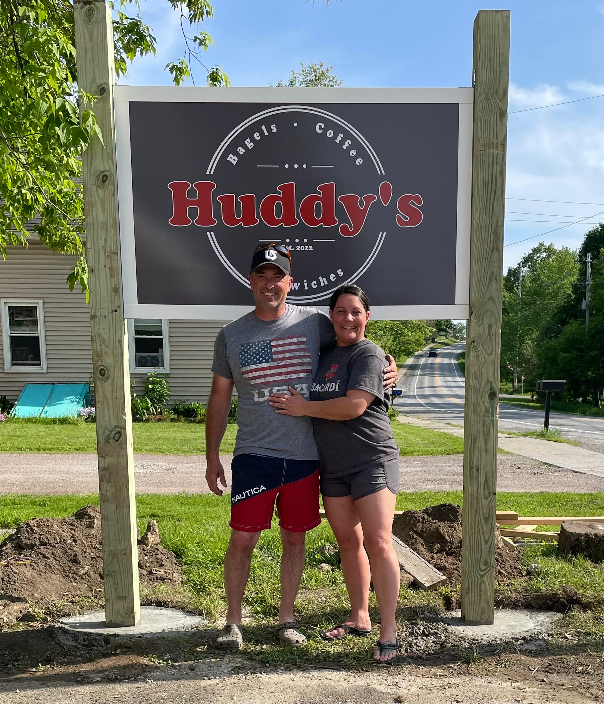 A couple stands proudly in front of a new restaurant sign that reads 'Huddy's' along a road, with greenery and construction materials in the background.