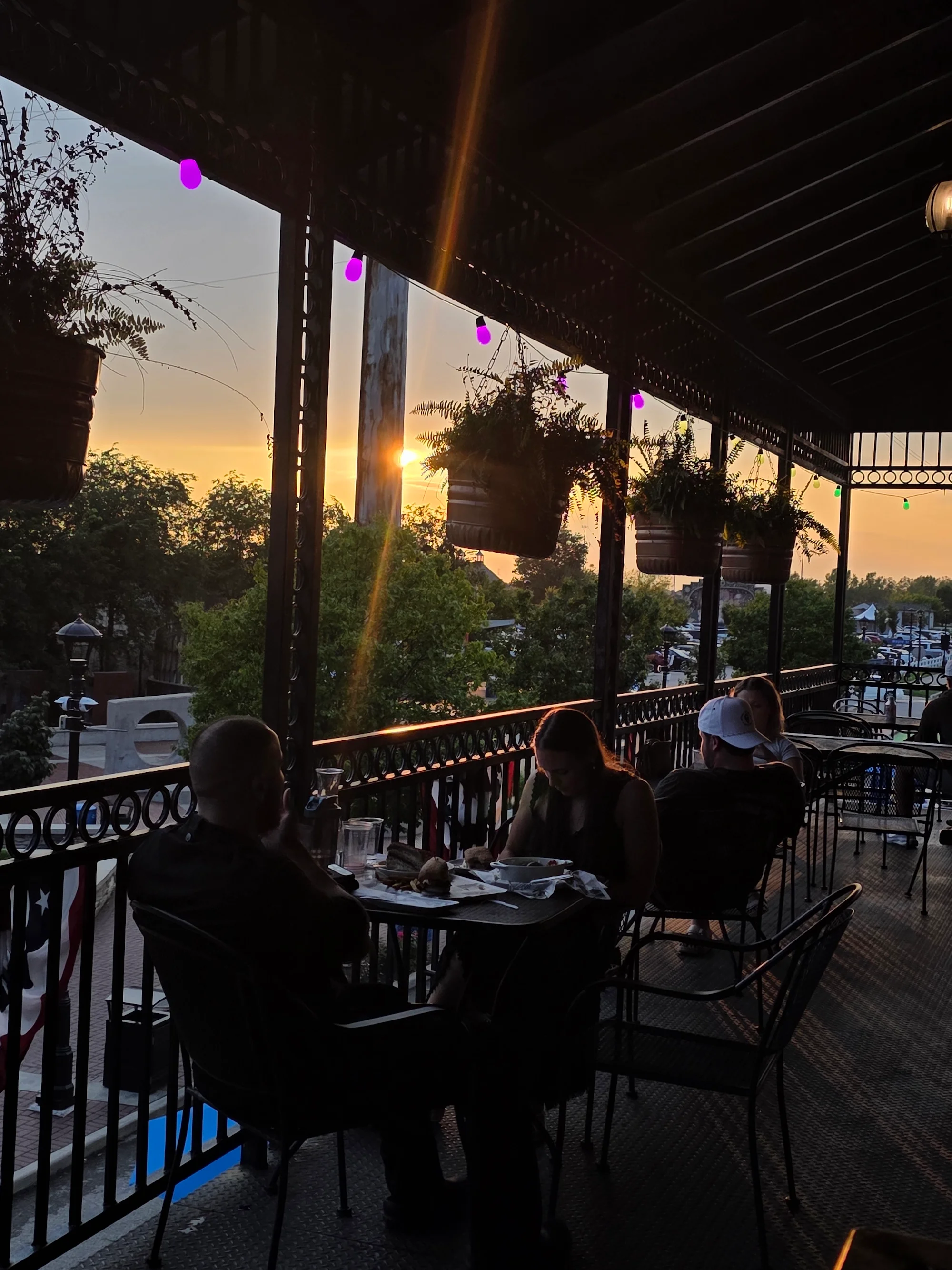 An outdoor dining area with a sunset in the background. The scene features hanging plants and warm lighting as people enjoy their meals.