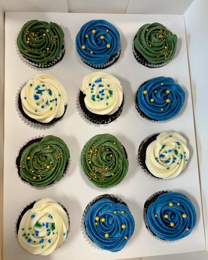 A collection of decorated cupcakes featuring blue, green, and white swirl frosting, adorned with colorful sprinkles and gold accents, arranged neatly in a white box.