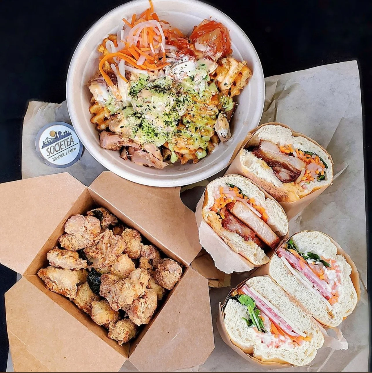 A variety of dishes including a bowl of loaded fries with chicken, pickled vegetables, and sauces, accompanied by three sandwiches and a box of fried pieces.