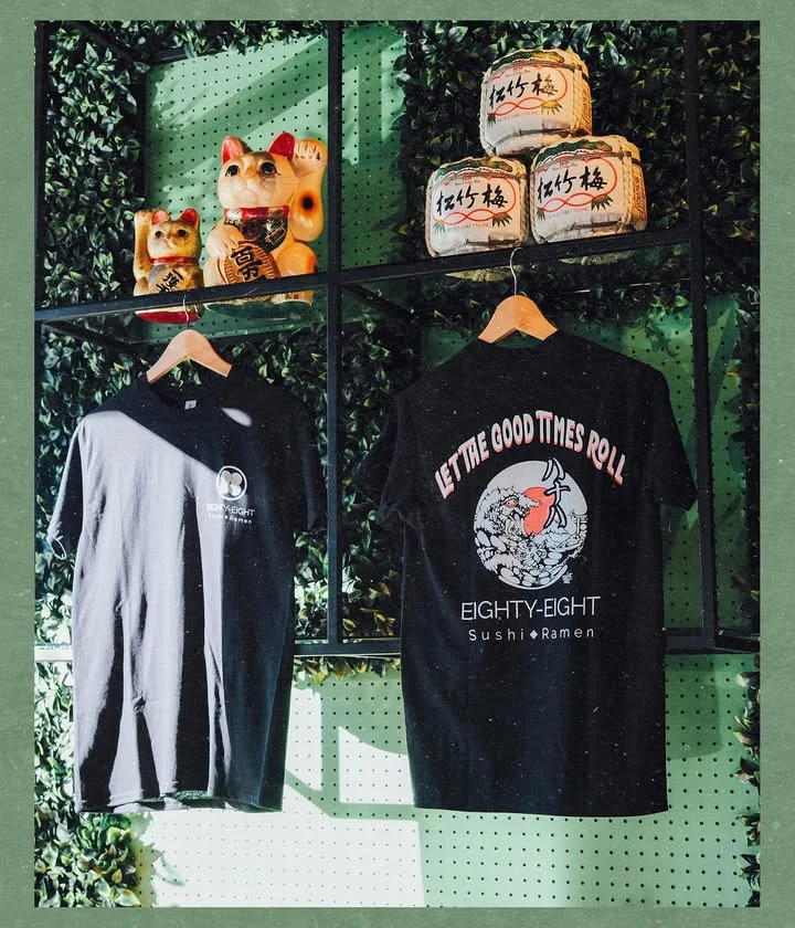 Two black t-shirts hanging on a display. One shirt features a graphic with the phrase 'LET THE GOOD TIMES ROLL' and the logo for a sushi and ramen brand. The second shirt has a simpler logo.