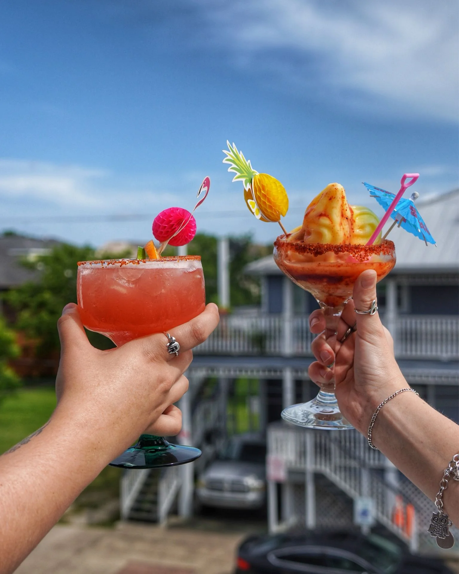 Two colorful cocktails with decorative garnishes are raised against a blue sky. One is a vibrant pink drink, while the other features a frozen fruit blend topped with an umbrella.