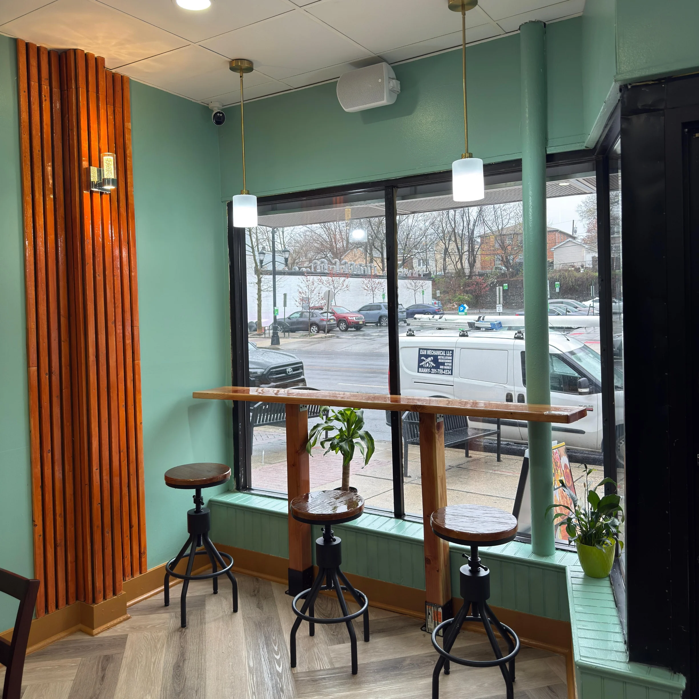 Three wooden stools at a café bar with teal walls, vertical wooden slats, and pendant lights. Outside the large windows, cars and a white van are visible on the street.