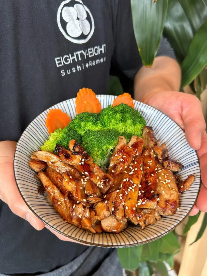 A bowl of grilled chicken garnished with sesame seeds, served with broccoli and carrot slices.