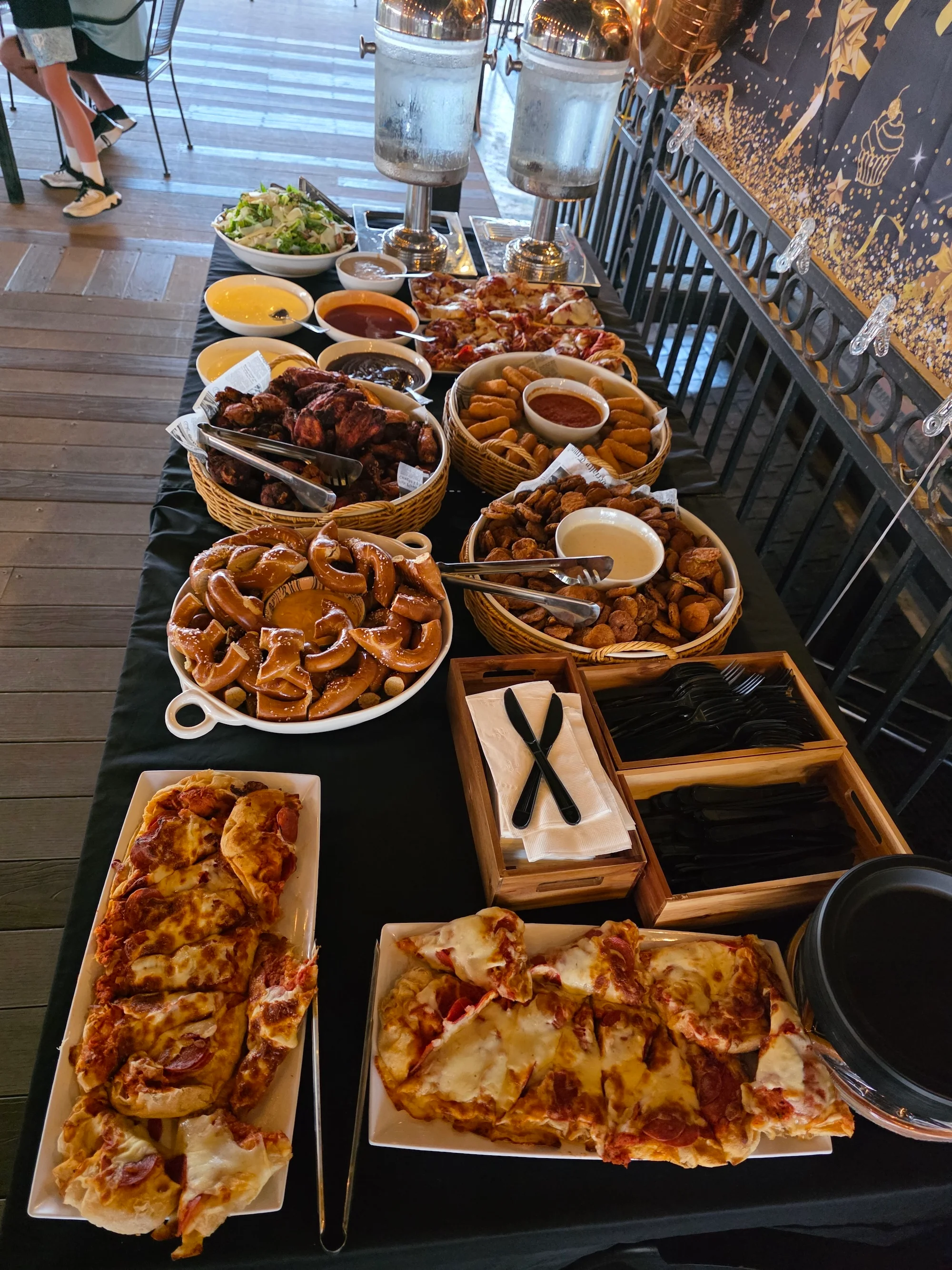 A bustling buffet spread with a variety of foods including salads, sauces, and pizzas, arranged on a black tablecloth. Two beverage dispensers are visible in the background.