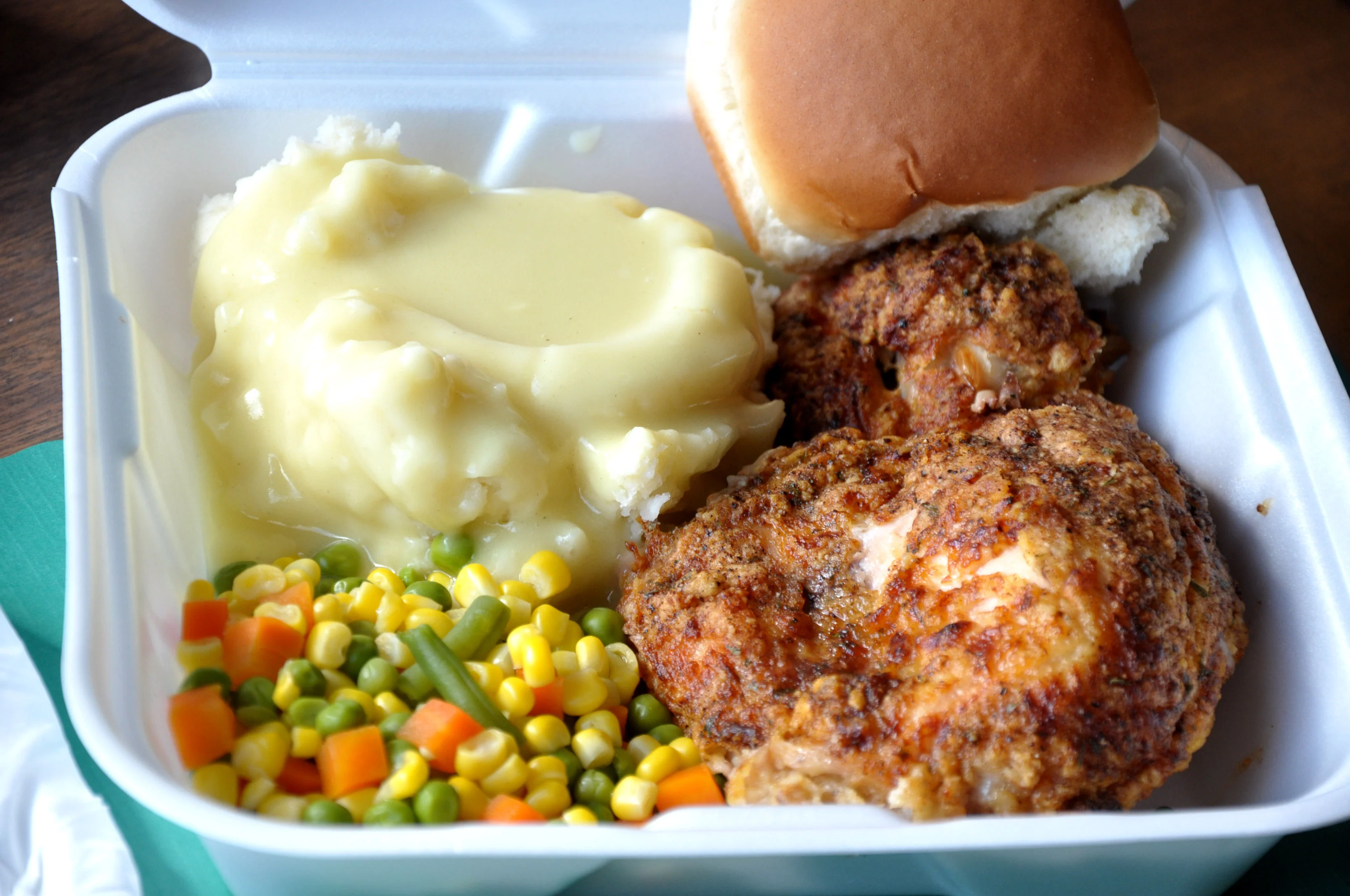 A takeout container featuring fried chicken, creamy mashed potatoes with gravy, and mixed vegetables including corn and carrots, along with a soft bun.