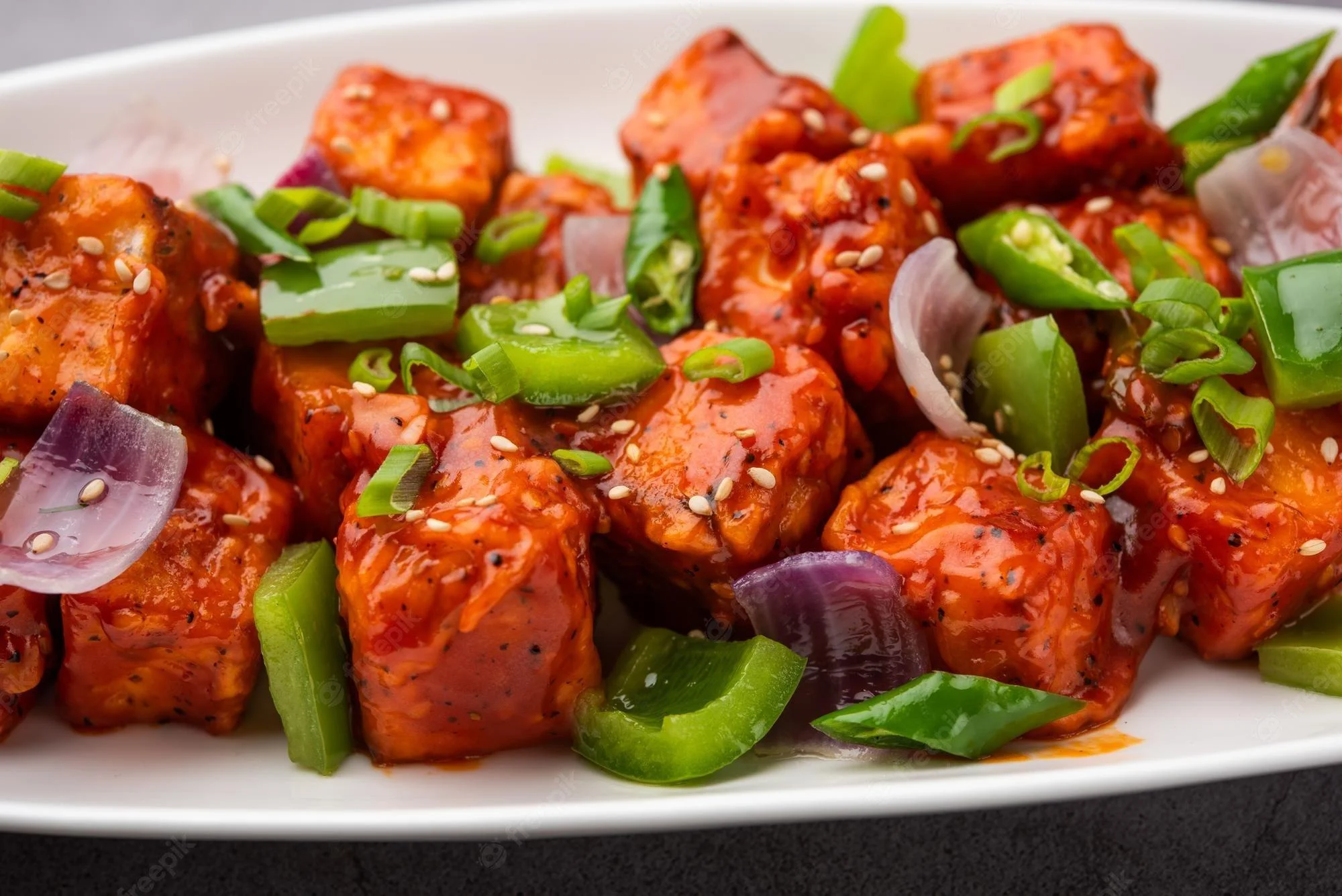 A plate of vibrant, red and orange cubes of fried tofu or paneer coated in a tangy sauce, mixed with diced green and purple bell peppers and garnished with chopped green onions.