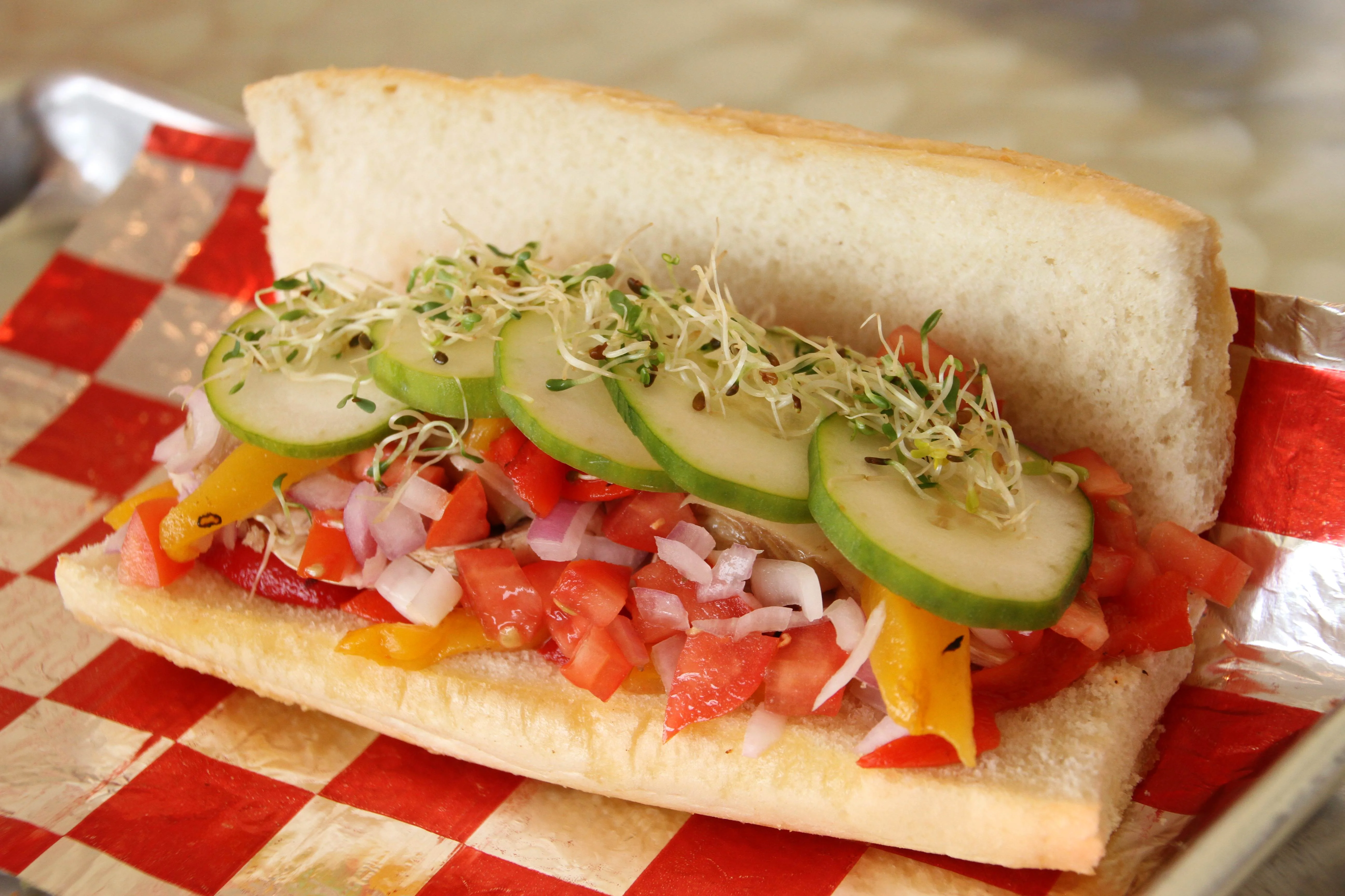 Fresh veggie sub sandwich on a hoagie roll with sliced cucumbers, alfalfa sprouts, diced tomatoes, onions, and colorful bell peppers, served on red and white checkered deli paper.