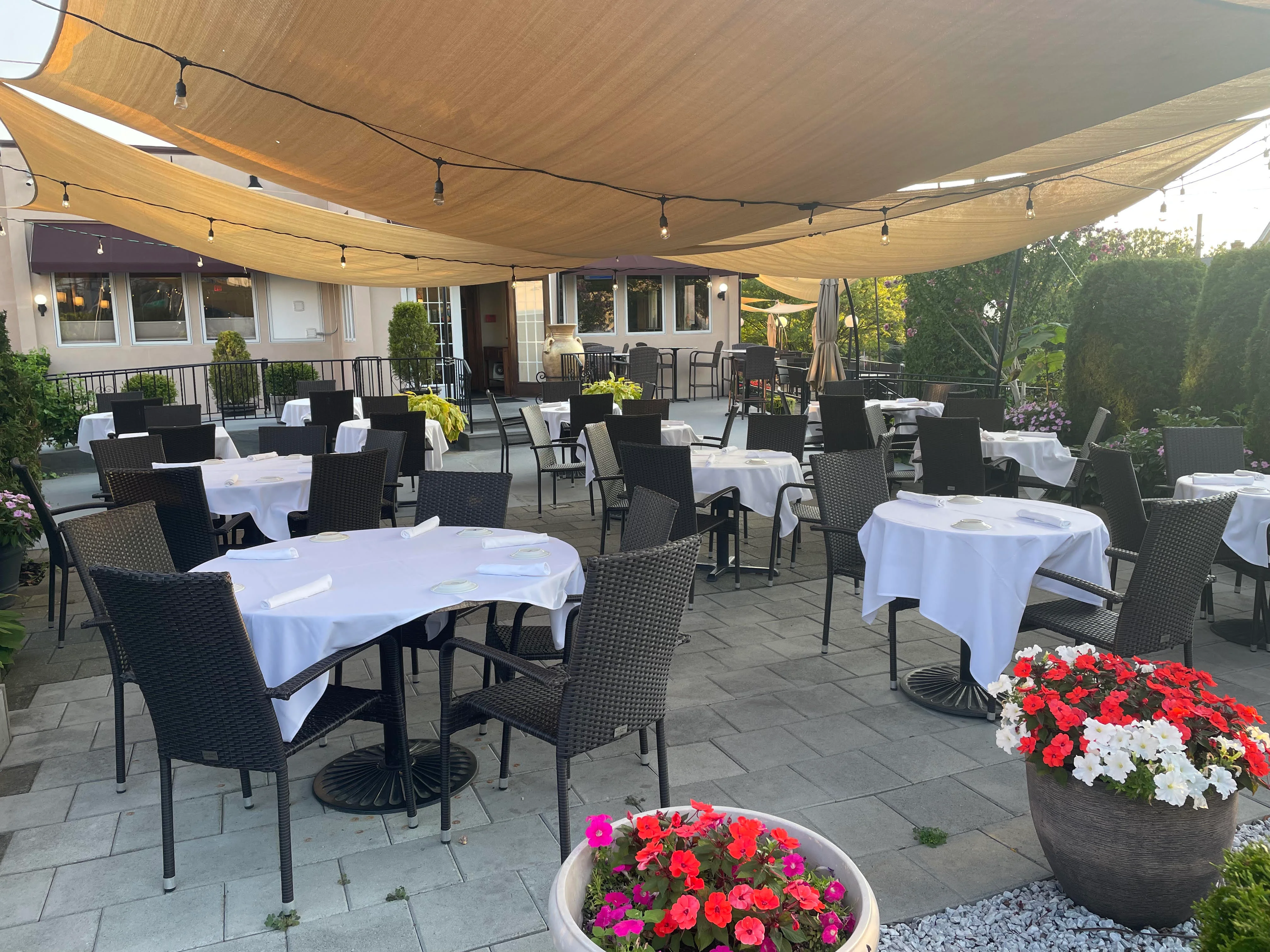 Toscana on Main patio with round tables covered in white cloths and surrounded by wicker chairs. Beige sunshade above and colorful flowers in planters