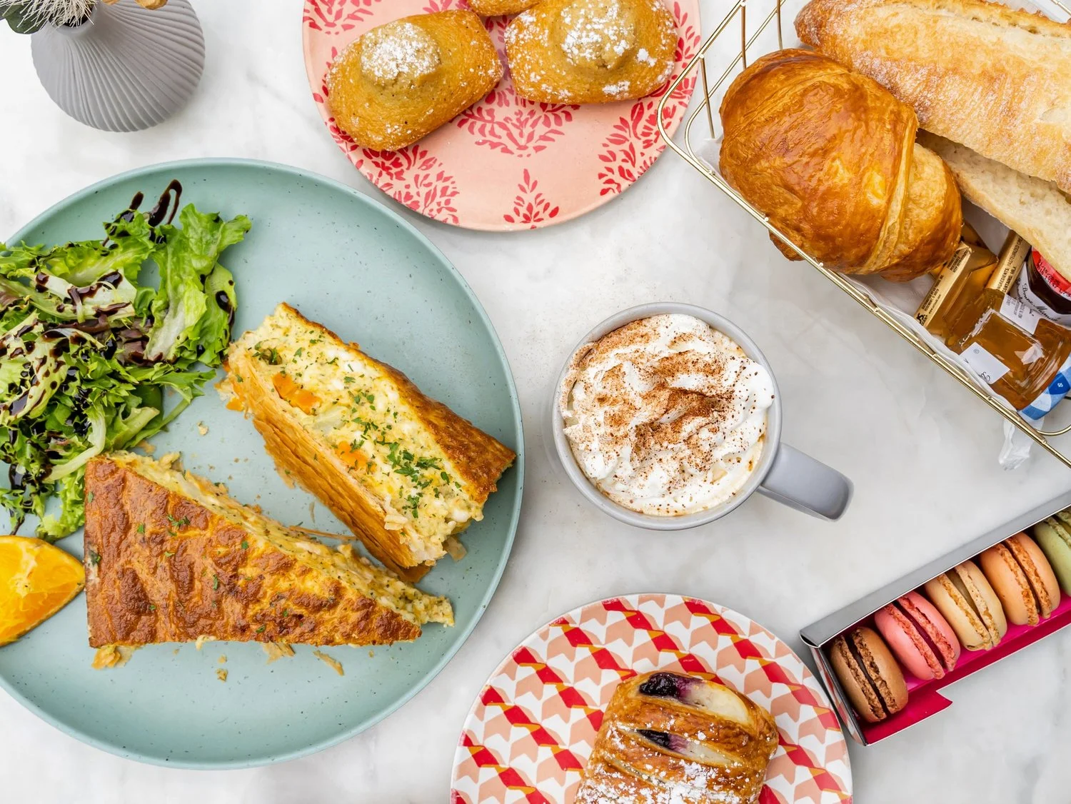 A delicious spread featuring a slice of savory quiche next to a fresh salad, accompanied by a mug of whipped cream topped beverage, a pastry, and an assortment of colorful macarons.