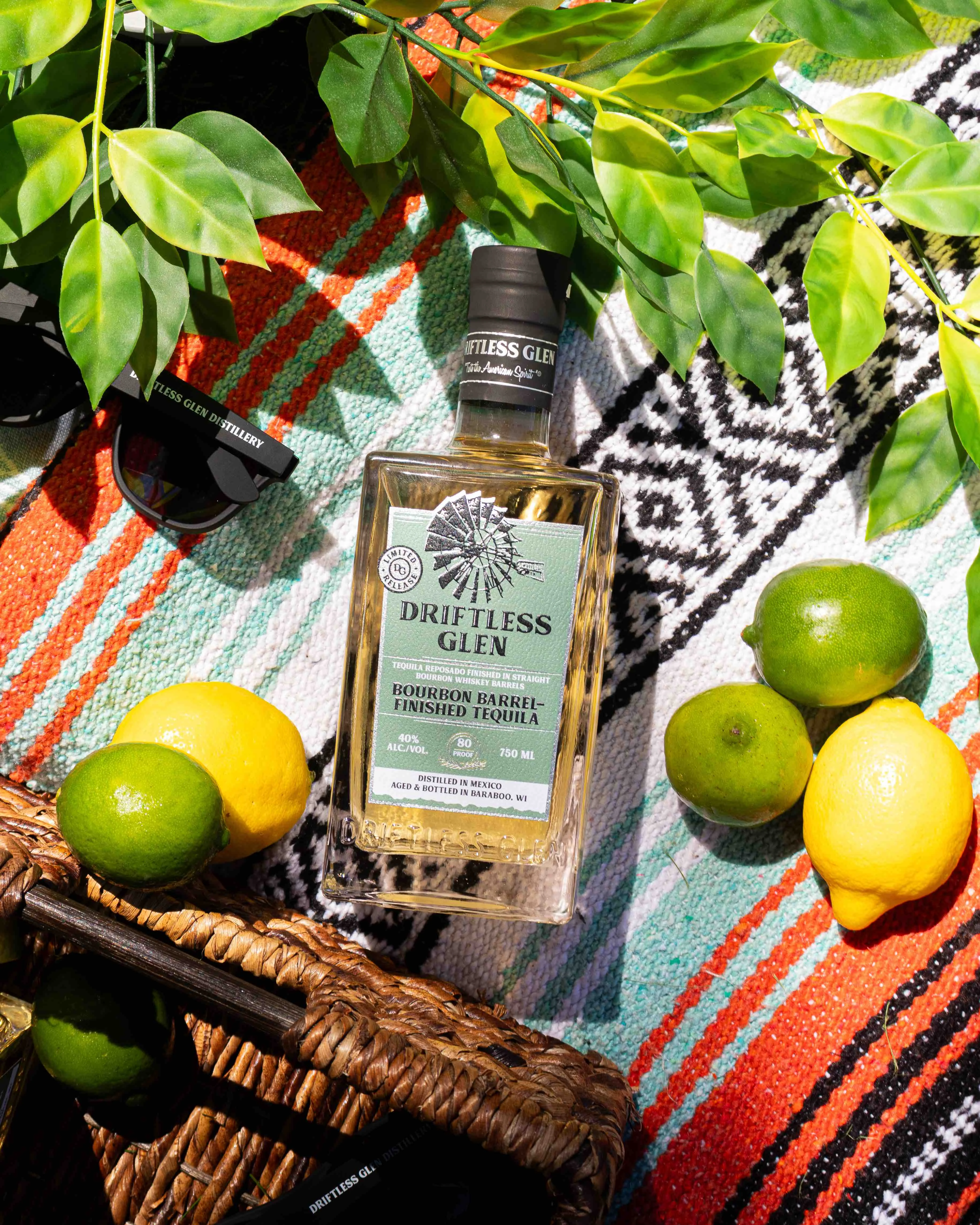 A bottle of Bourbon Barrel-Finished Tequila with some limes, lemons, and sunglasses.
