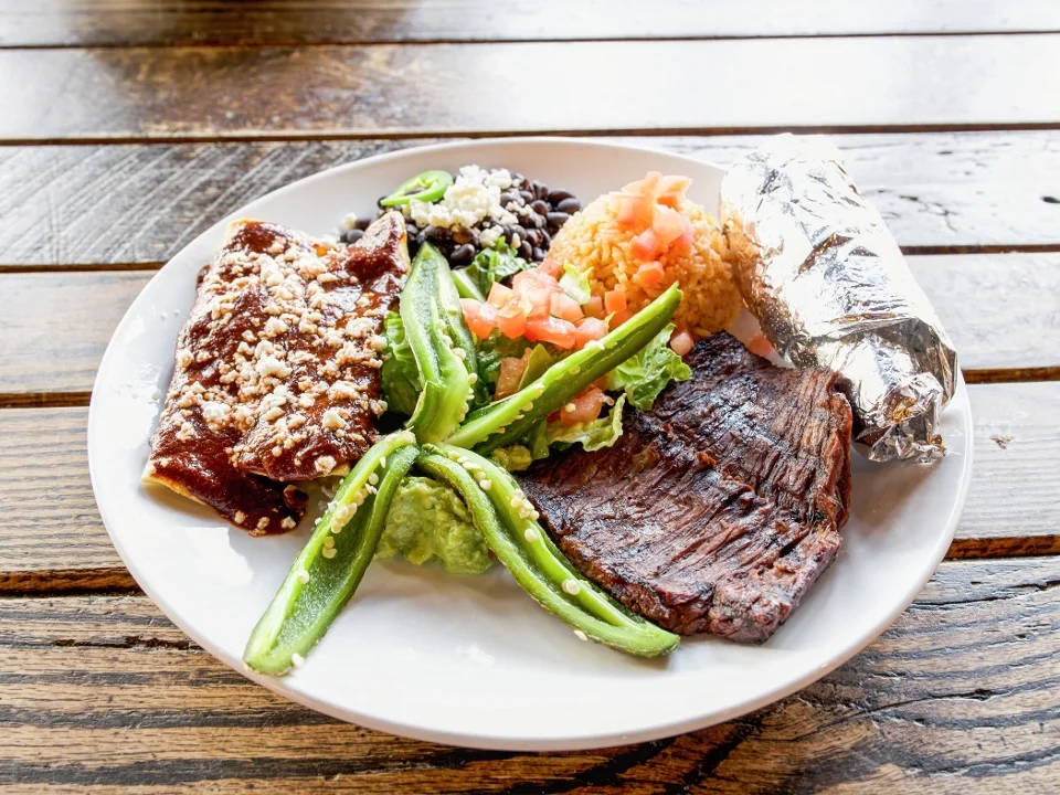 A hearty plate of Mexican food featuring grilled meat, enchiladas, guacamole, beans, and a side of rice.