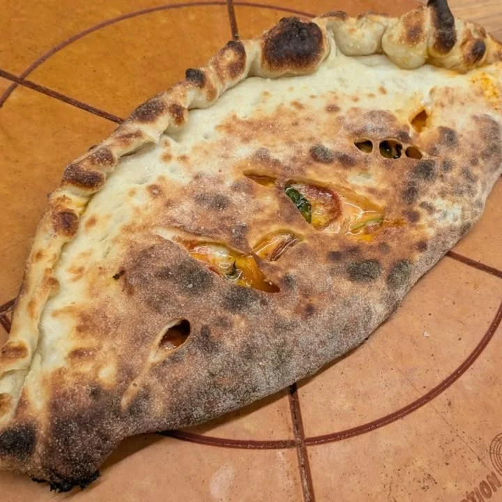 A calzone with a slightly charred crust, showing some filling peeking through the openings. It is placed on a wooden surface.