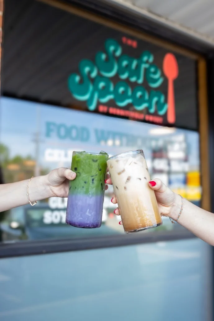 Two colorful drinks held up in toast, one green and the other creamy, in front of the entrance of a shop. The background features a sign with vibrant text.