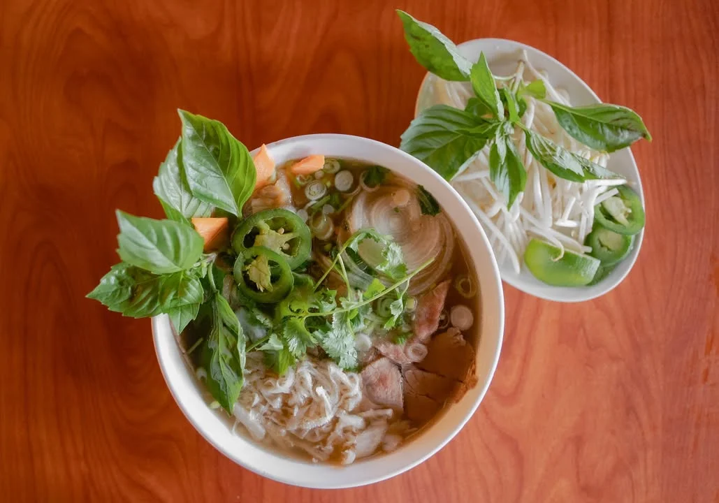 A bowl of noodle soup topped with fresh herbs, sliced meat, and vegetables. A side plate contains additional herbs and lime.