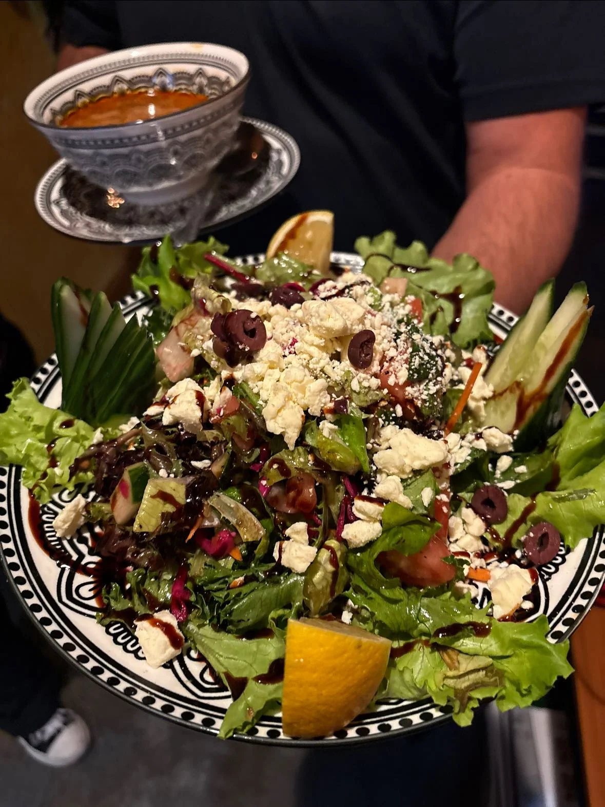A vibrant salad topped with olives, cheese, and citrus, served on a beautifully patterned plate. A bowl of soup accompanies it.