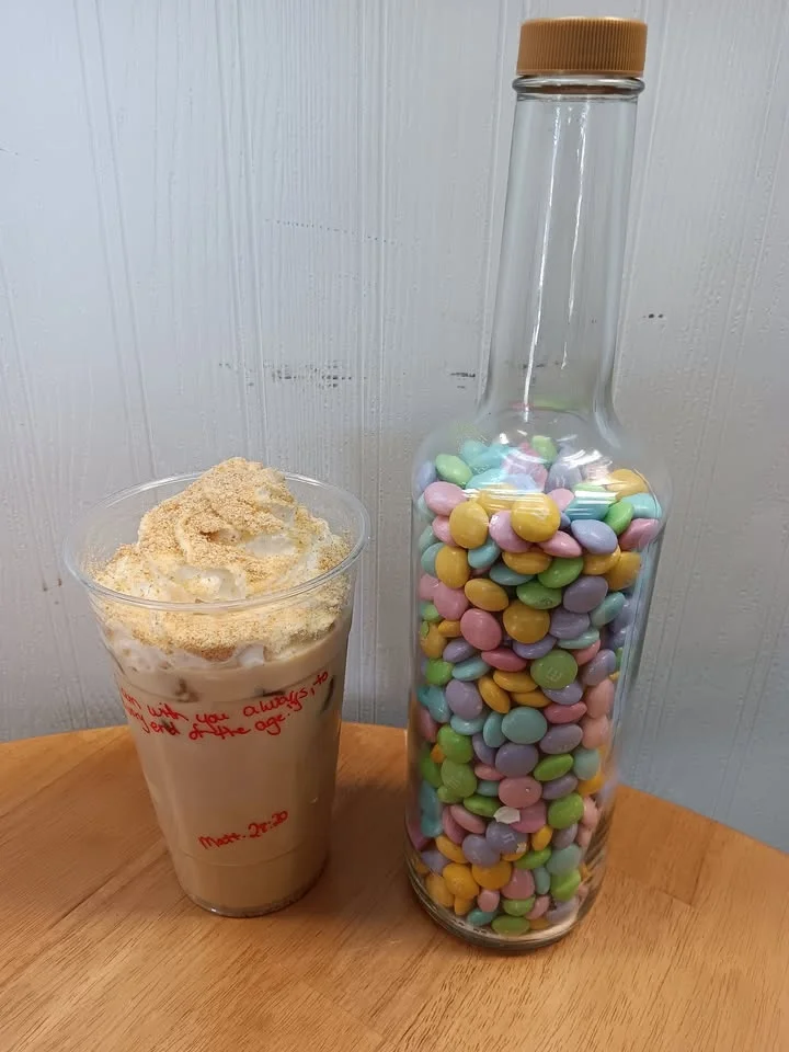 A refreshing beverage topped with whipped cream, accompanied by a glass jar filled with colorful candy, placed on a wooden table.