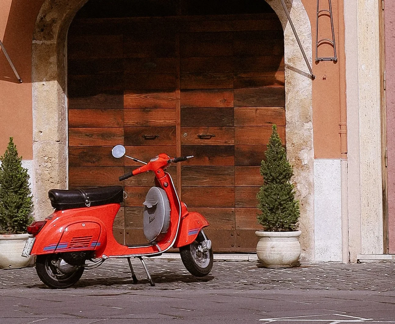 Red Vespa parked on cobblestones in front of an arched wooden doorway in Rome, featured by Daje Pinsa Roman Pizza in Orlando.