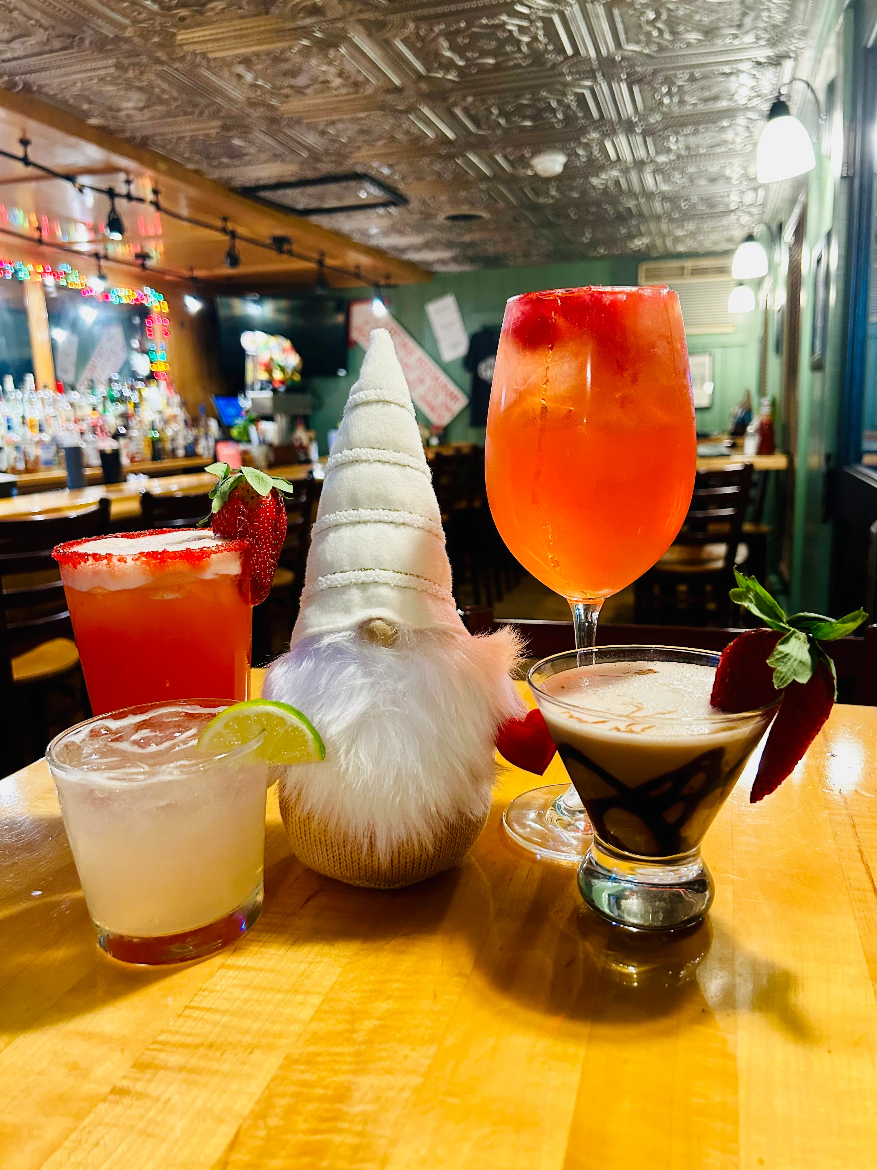 A selection of colorful cocktails on a wooden table, featuring a whimsical gnome figure among the drinks.