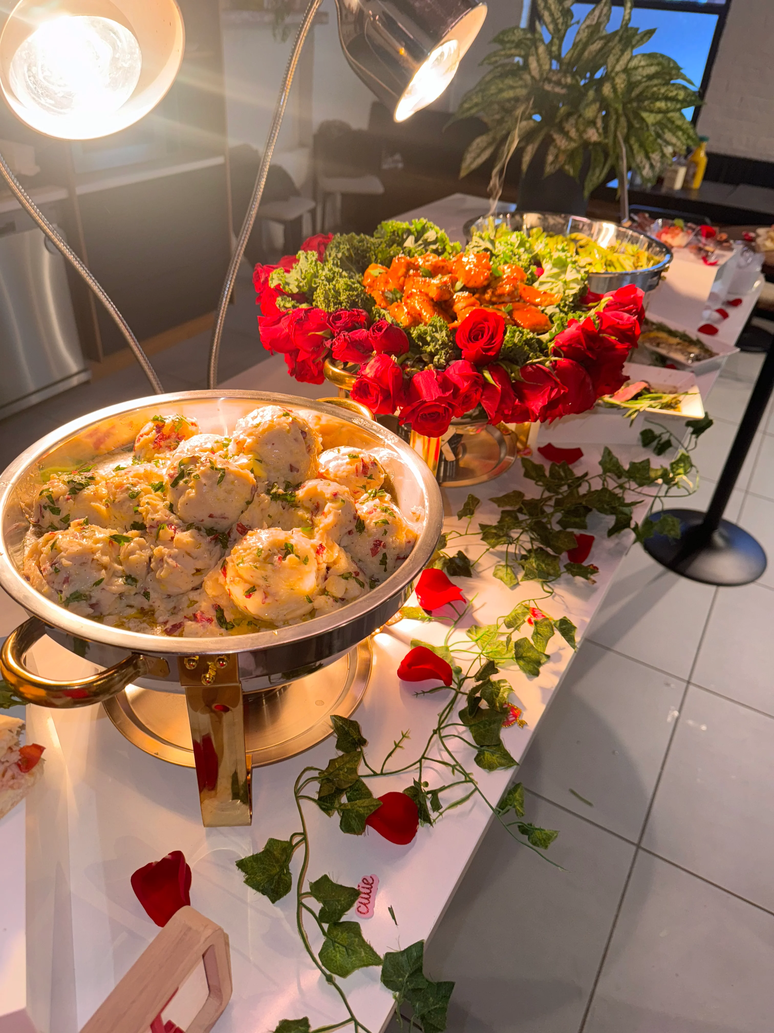 A beautifully arranged buffet featuring platters of food under warm lighting. One dish contains round servings of creamy food garnished with herbs, while another showcases bright orange treats nestled among fresh green lettuce and vibrant roses.