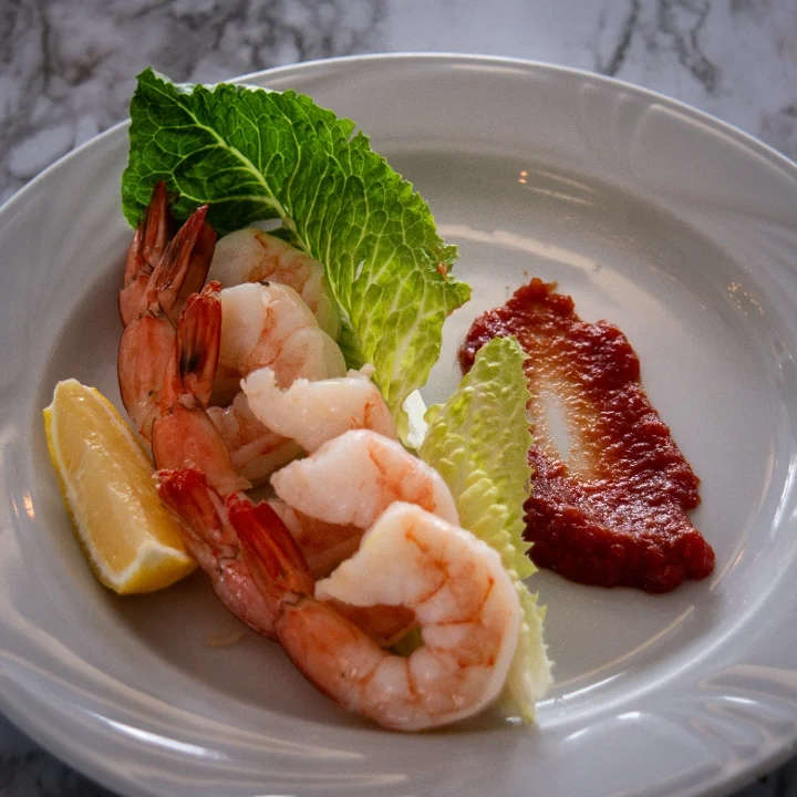A plate of shrimp served with lettuce leaves, a slice of lemon, and a side of sauce. The dish is arranged artistically on a white plate.